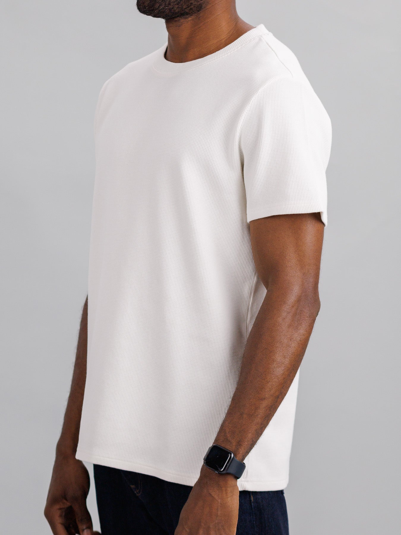 A person, wearing the ID Menswear Urban Waffle Knit Tee in a white cotton-spandex blend and a smartwatch, stands against a neutral background. The casual T-shirt design is visible from the neck down.