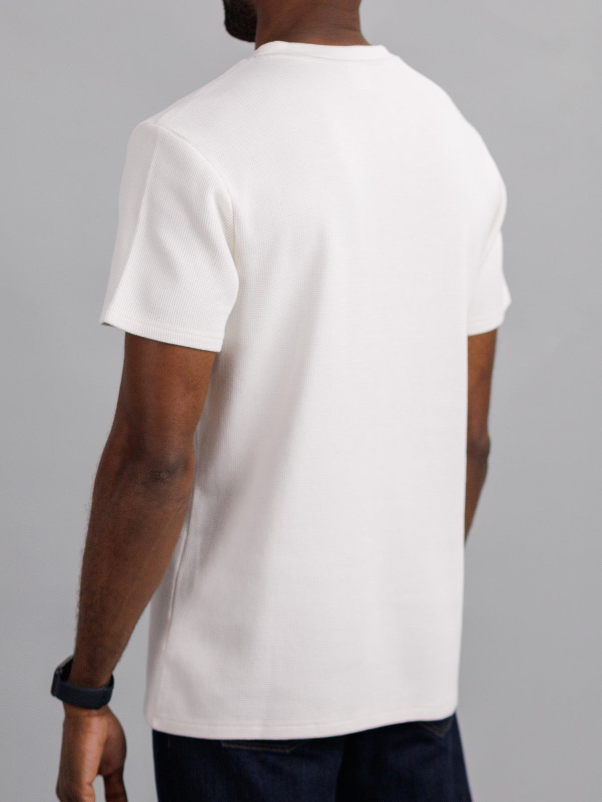 A person stands with their back to the camera, wearing an Urban Waffle Knit Tee by ID Menswear and dark pants. A smartwatch on their left wrist complements the casual attire against a simple gray background.