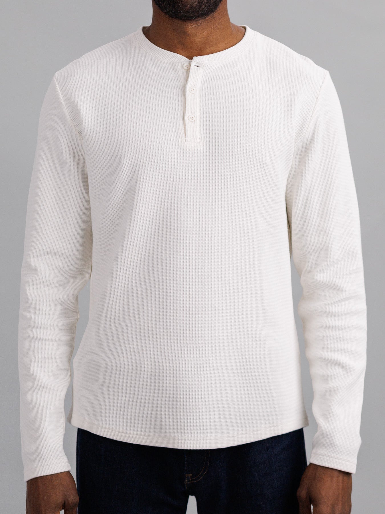 A person dons the ID Menswear Freestyle Waffle Knit Henley, a regular fit, long-sleeve shirt made from a soft cotton-spandex blend. It features a round neckline and short button placket while they stand against a gray backdrop, with part of their jeans visible.