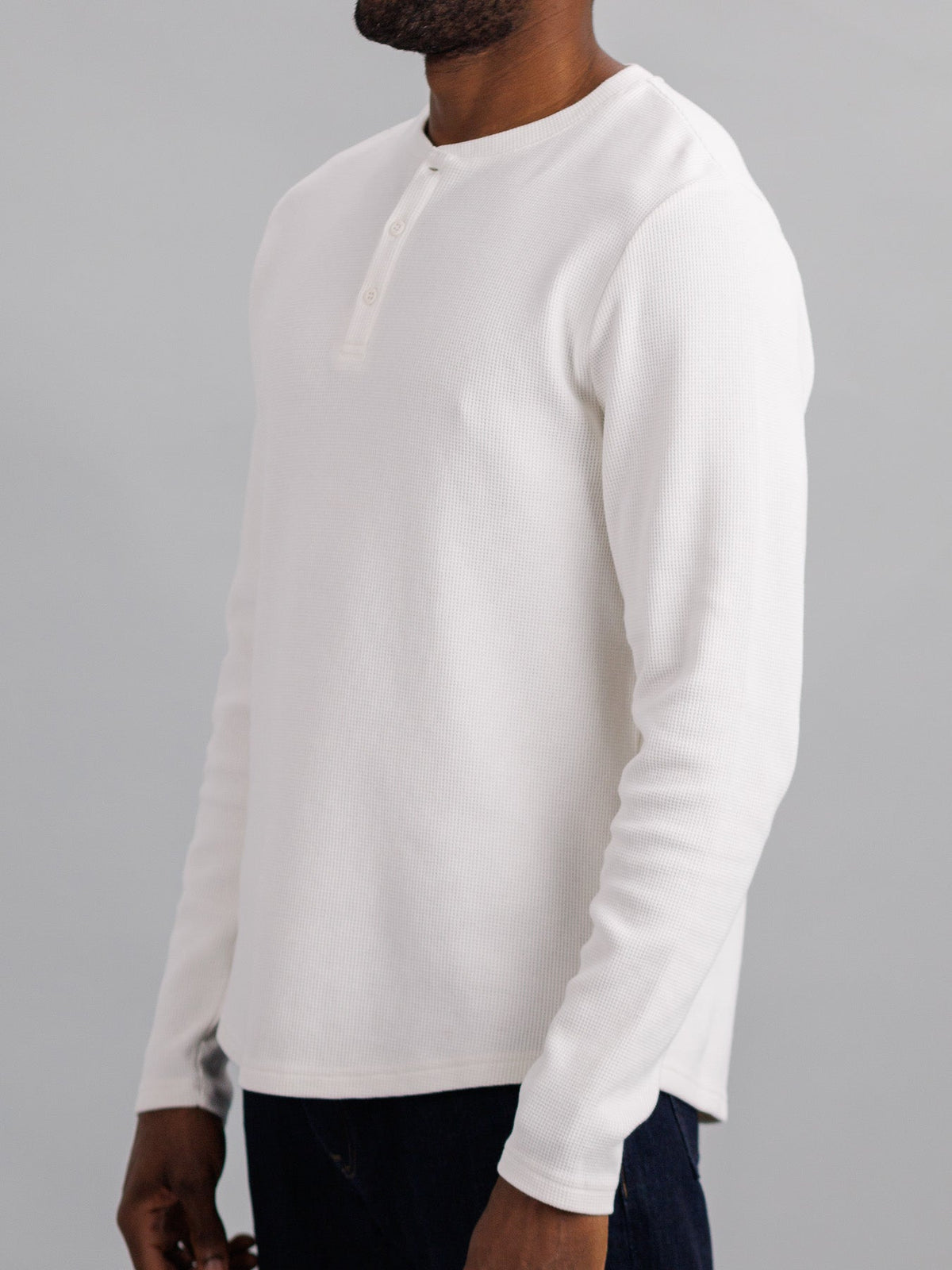 Someone wears the ID Menswear Freestyle Waffle Knit Henley—a white, waffle-knitted shirt with buttons—against a gray background, facing slightly sideways. The cotton-spandex blend casual top is paired with dark pants, focusing on the upper body.