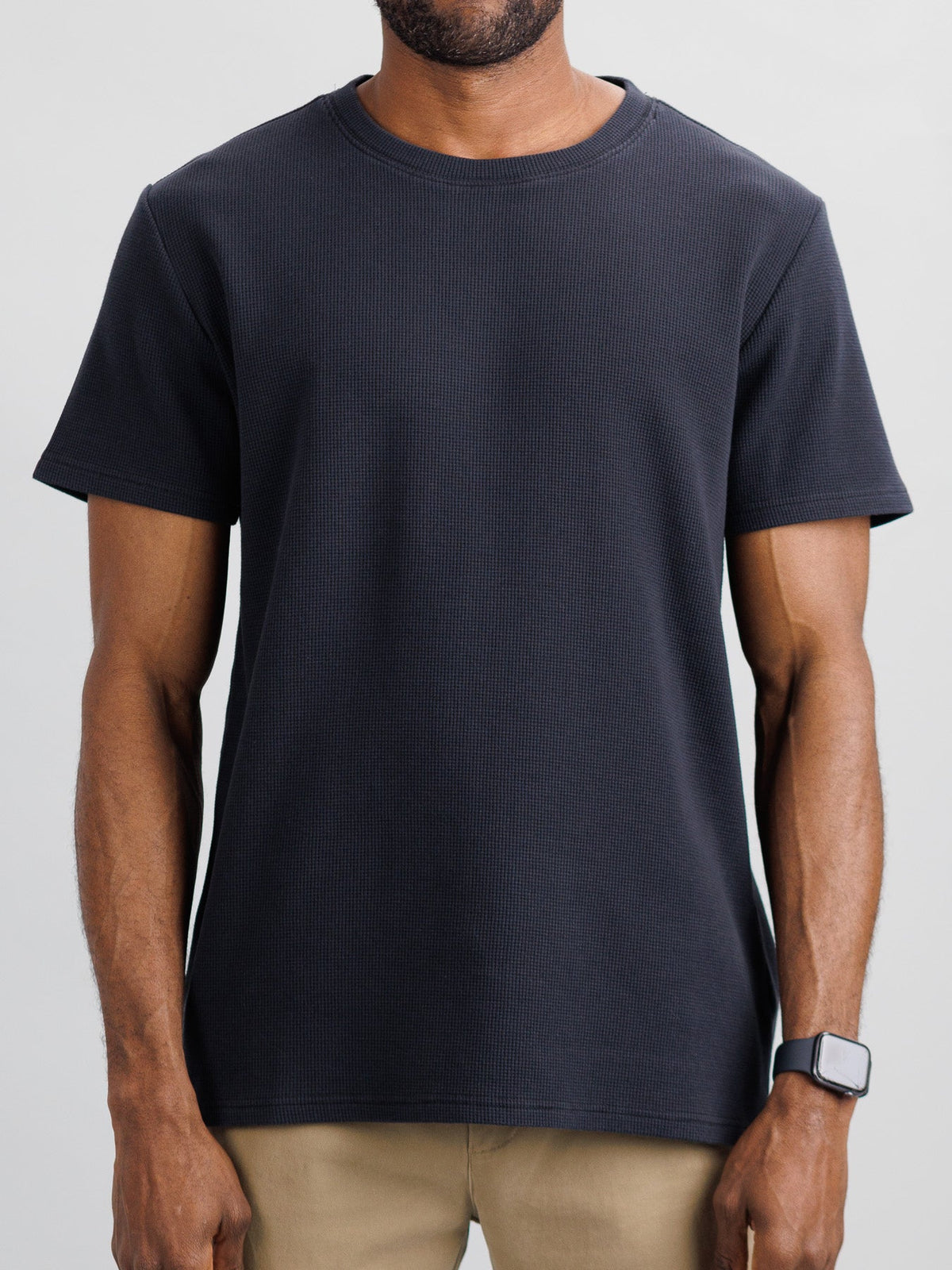 A man in an ID Menswear Urban Waffle Knit Tee and beige pants stands against a neutral background, exuding effortless style. A smartwatch on his left wrist adds a modern touch to his casual look.