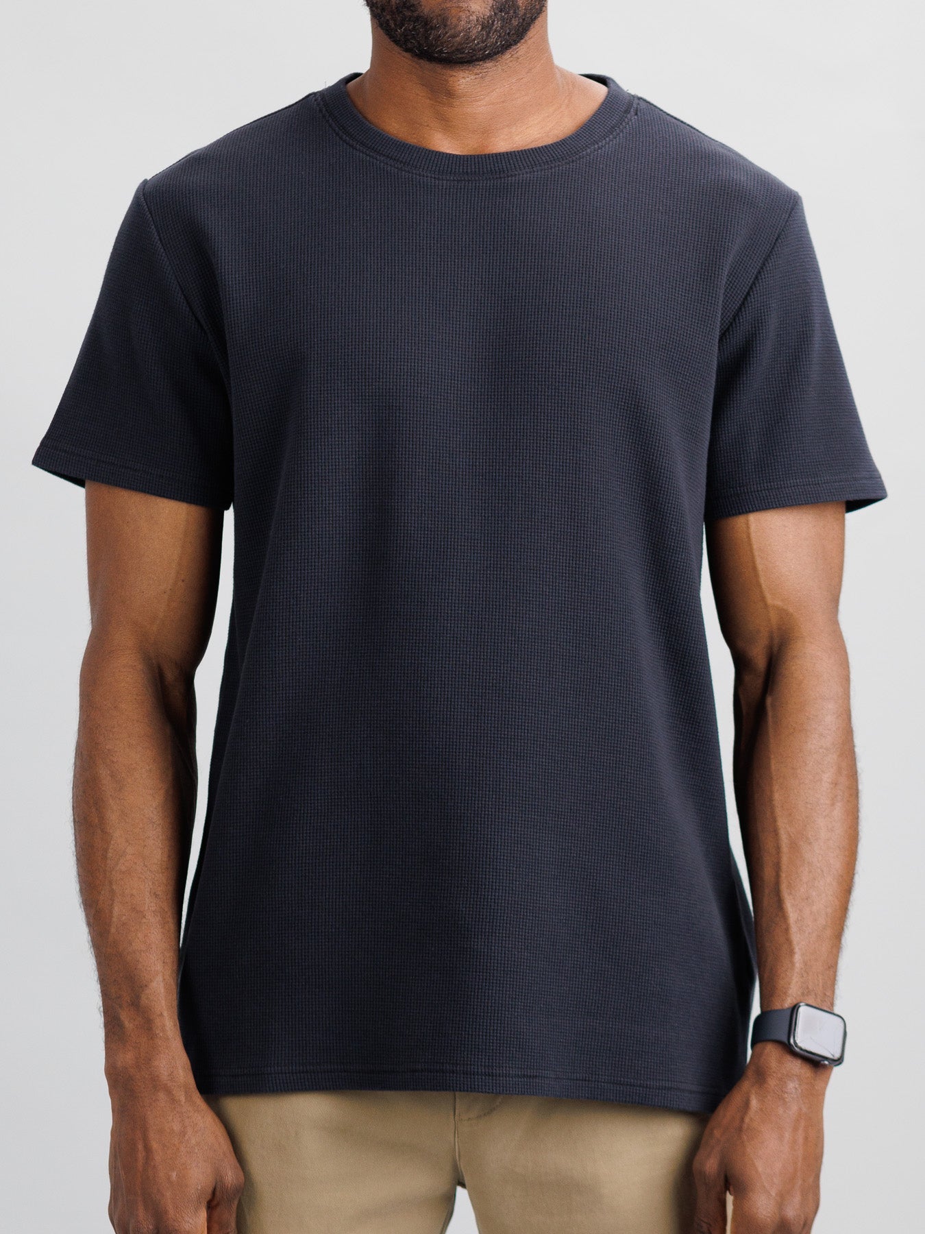 A man in an ID Menswear Urban Waffle Knit Tee and beige pants stands against a neutral background, exuding effortless style. A smartwatch on his left wrist adds a modern touch to his casual look.