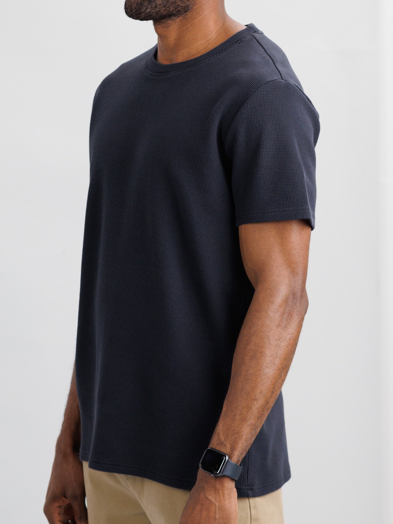 Wearing ID Menswears Urban Waffle Knit Tee in black paired with beige pants, someone stands against a neutral background. The ribbed texture of the tee adds sophistication, while a smartwatch gleams on their left wrist, completing the stylish yet comfortable look.