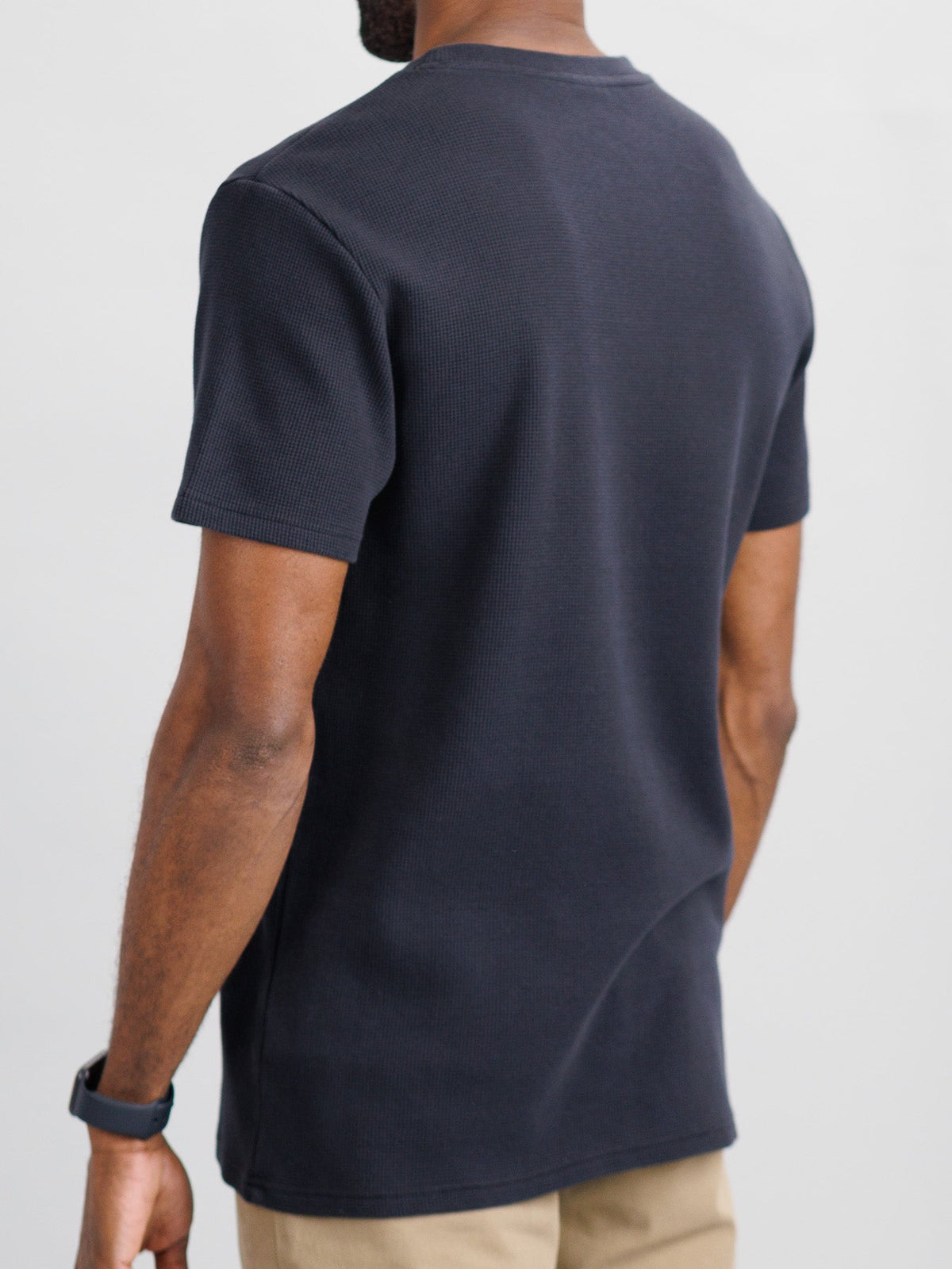 A person in casual wear stands with their back to the camera, wearing ID Menswears Urban Waffle Knit Tee in a plain dark color and beige pants. The soft cotton-spandex blend ensures a comfortable fit. A watch is on their left wrist against a neutral, light background.