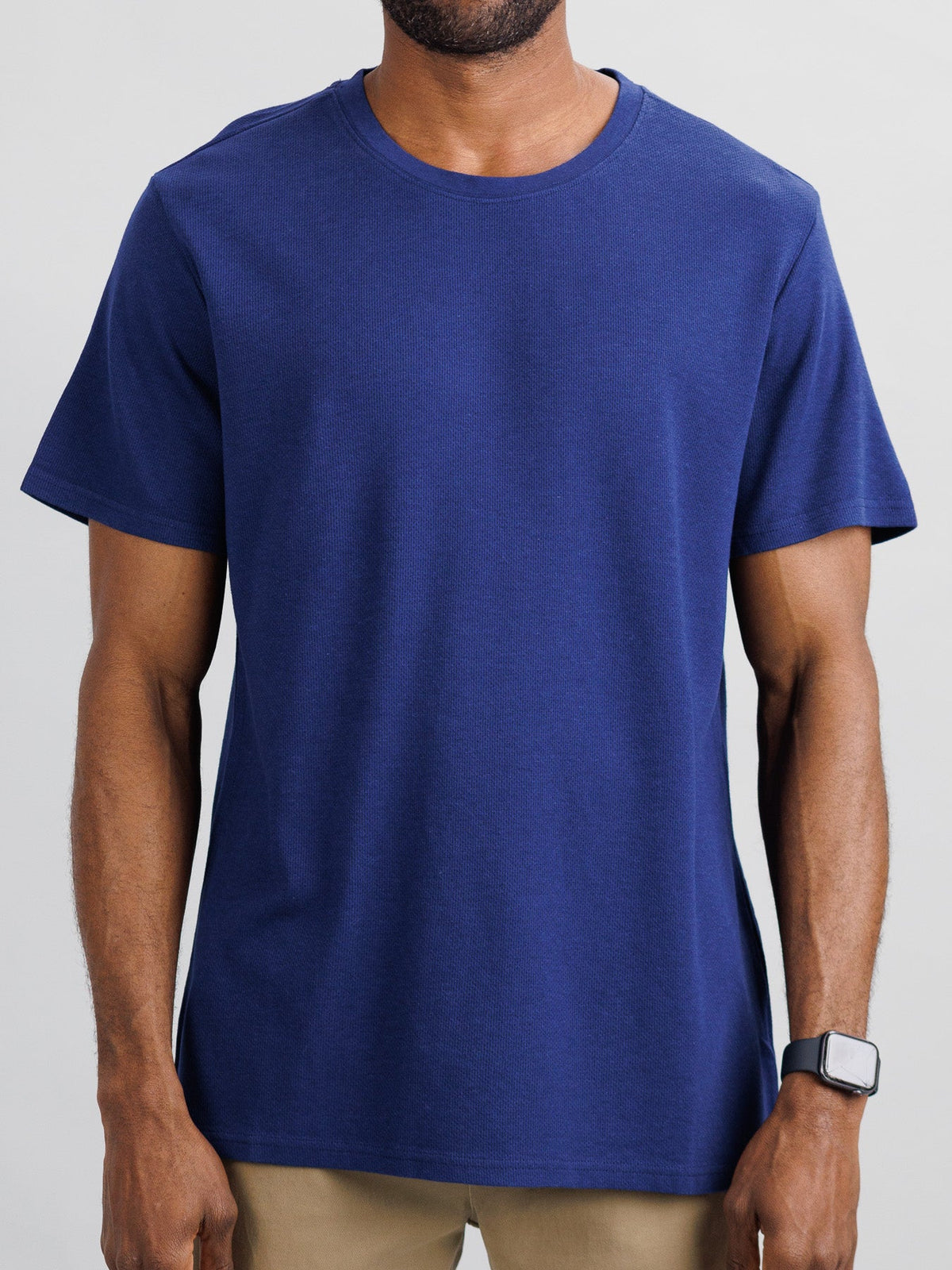 A man models the Urban Honeycomb Mesh Tee by ID Menswear in plain blue and beige pants against a light gray background, highlighting its bamboo-organic cotton blend for breathability. His smartwatch is visible, but his face isnt shown as the focus is on his torso and arms.