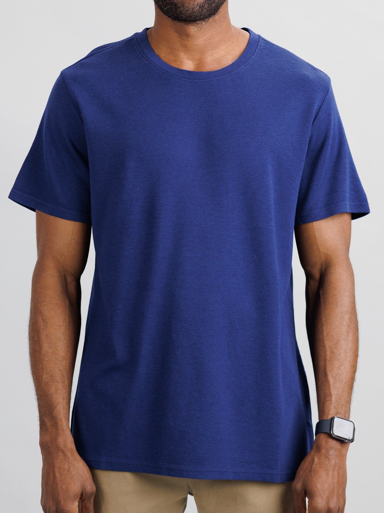 A man models the Urban Honeycomb Mesh Tee by ID Menswear in plain blue and beige pants against a light gray background, highlighting its bamboo-organic cotton blend for breathability. His smartwatch is visible, but his face isnt shown as the focus is on his torso and arms.