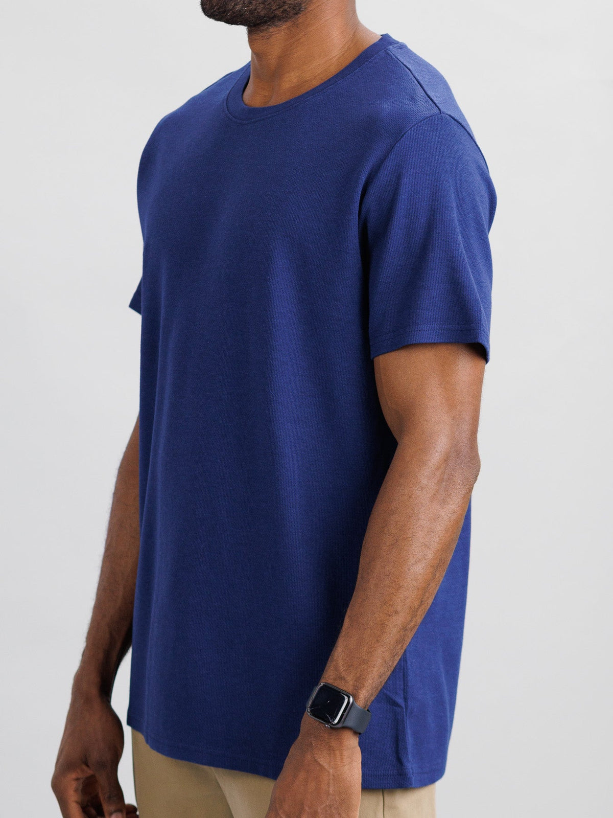 A person stands with relaxed arms, wearing ID Menswears Urban Honeycomb Mesh Tee in plain blue and beige pants. They have a watch on their left wrist against a neutral, light gray background.