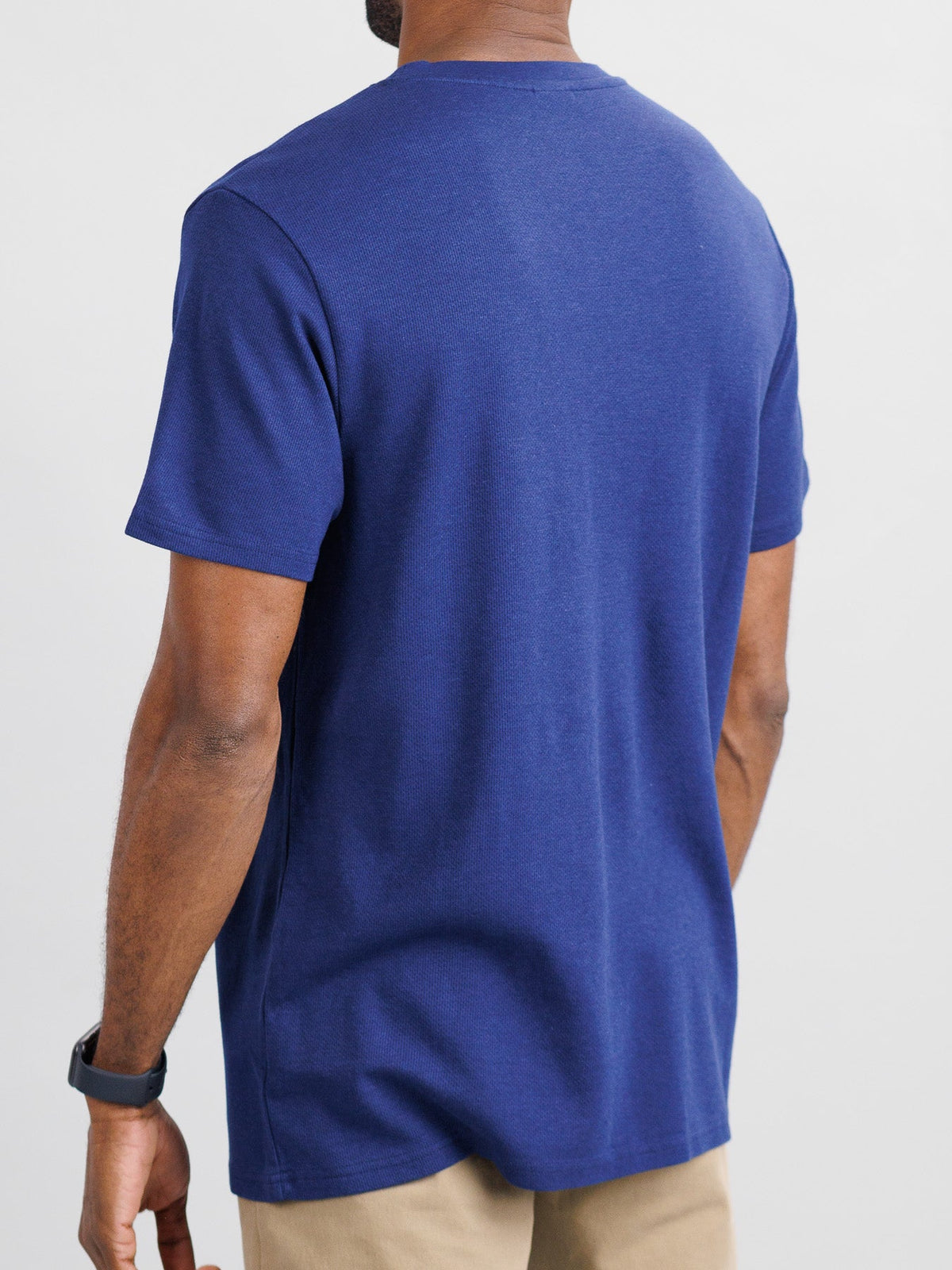 A person is seen from behind wearing ID Menswears Urban Honeycomb Mesh Tee in navy blue and beige pants. Their left arm is bent, displaying a black wristwatch. The plain white background highlights the breathable texture of their apparel.