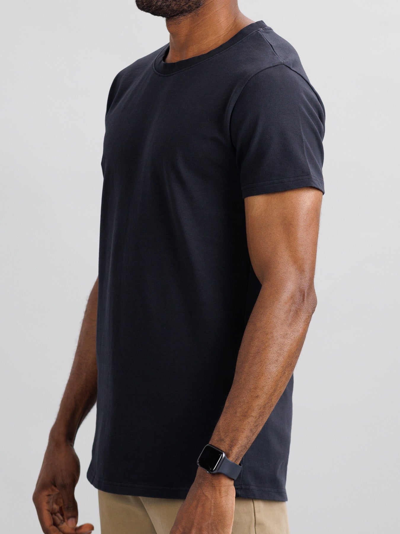 A person in a slim fit City Organic Cotton Tee by ID Menswear and beige pants is seen from the side. The black T-shirt enhances the look of their smartwatch on the left wrist. The background is plain and neutral.