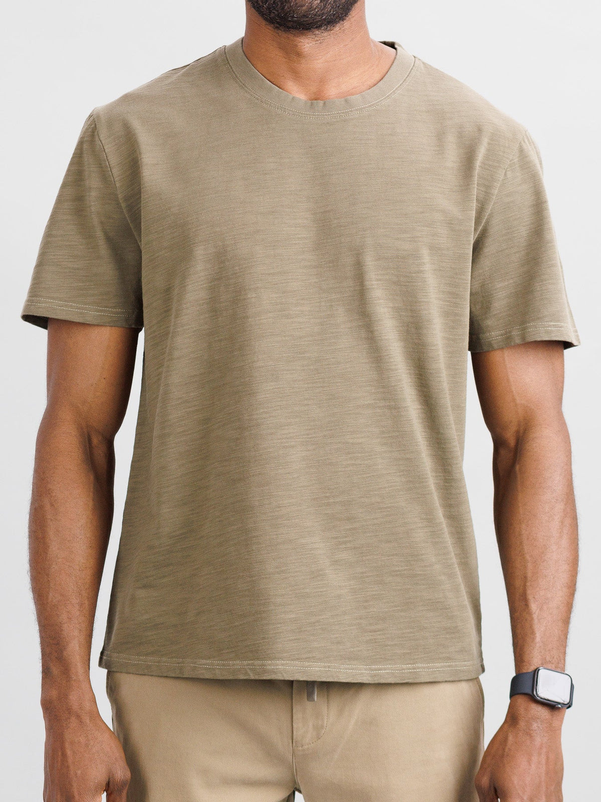 A man in an ID Menswear Big Flip Slub Yarn Cotton Tee and matching pants, both in plain olive green, stands against a light background. The 100% cotton fabric features a boxy fit. He wears a dark watch on his left wrist with his hands relaxed at his sides.