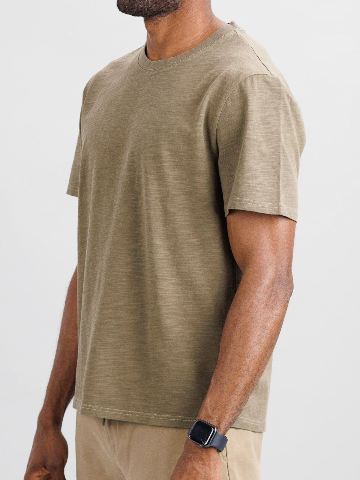 Someone stands against a neutral background wearing the ID Menswear Big Flip Slub Yarn Cotton Tee in a boxy fit, beige pants, and a smartwatch on their left wrist. The image focuses on the casual 100% cotton outfit, cropped at the neck and knees.