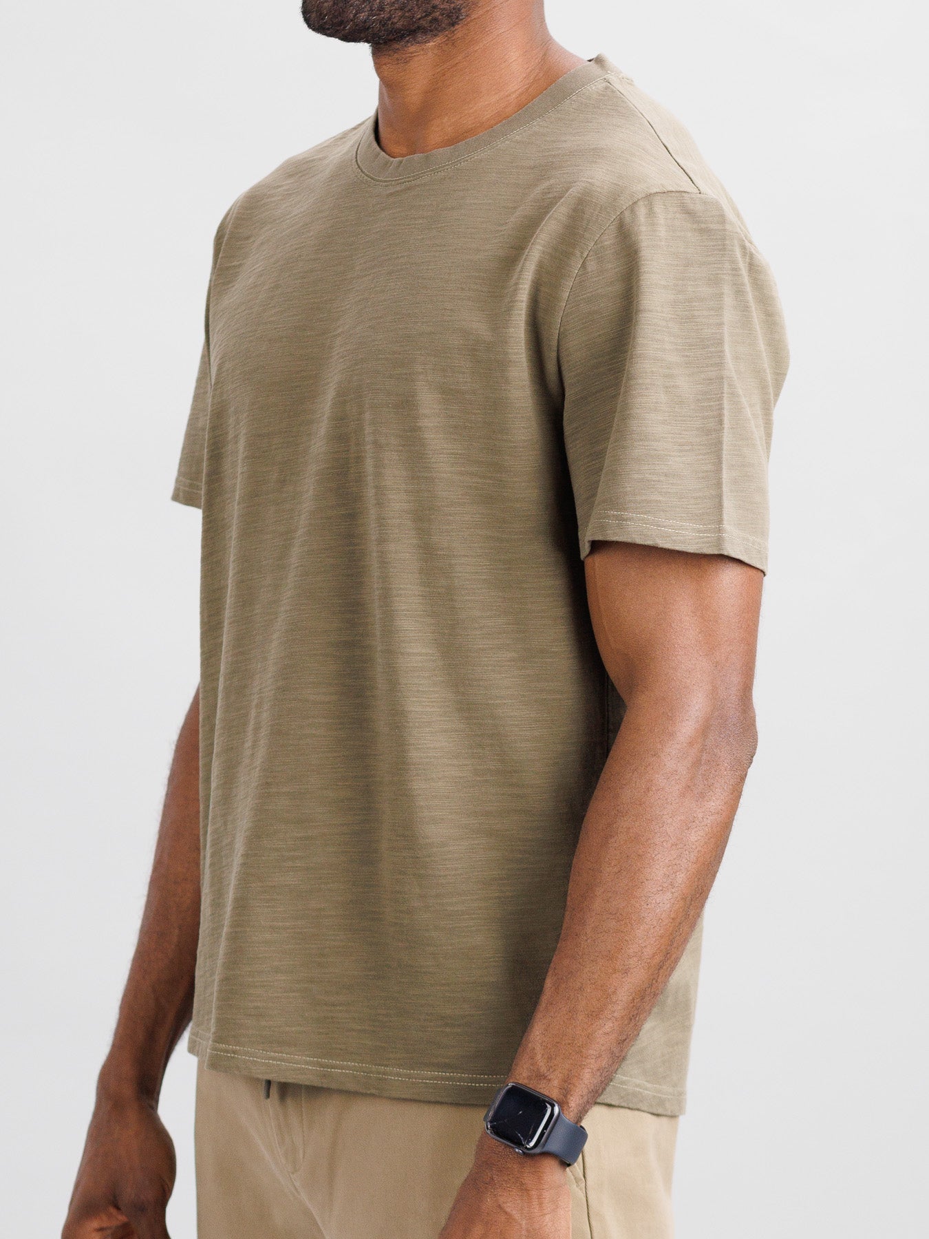 Someone stands against a neutral background wearing the ID Menswear Big Flip Slub Yarn Cotton Tee in a boxy fit, beige pants, and a smartwatch on their left wrist. The image focuses on the casual 100% cotton outfit, cropped at the neck and knees.