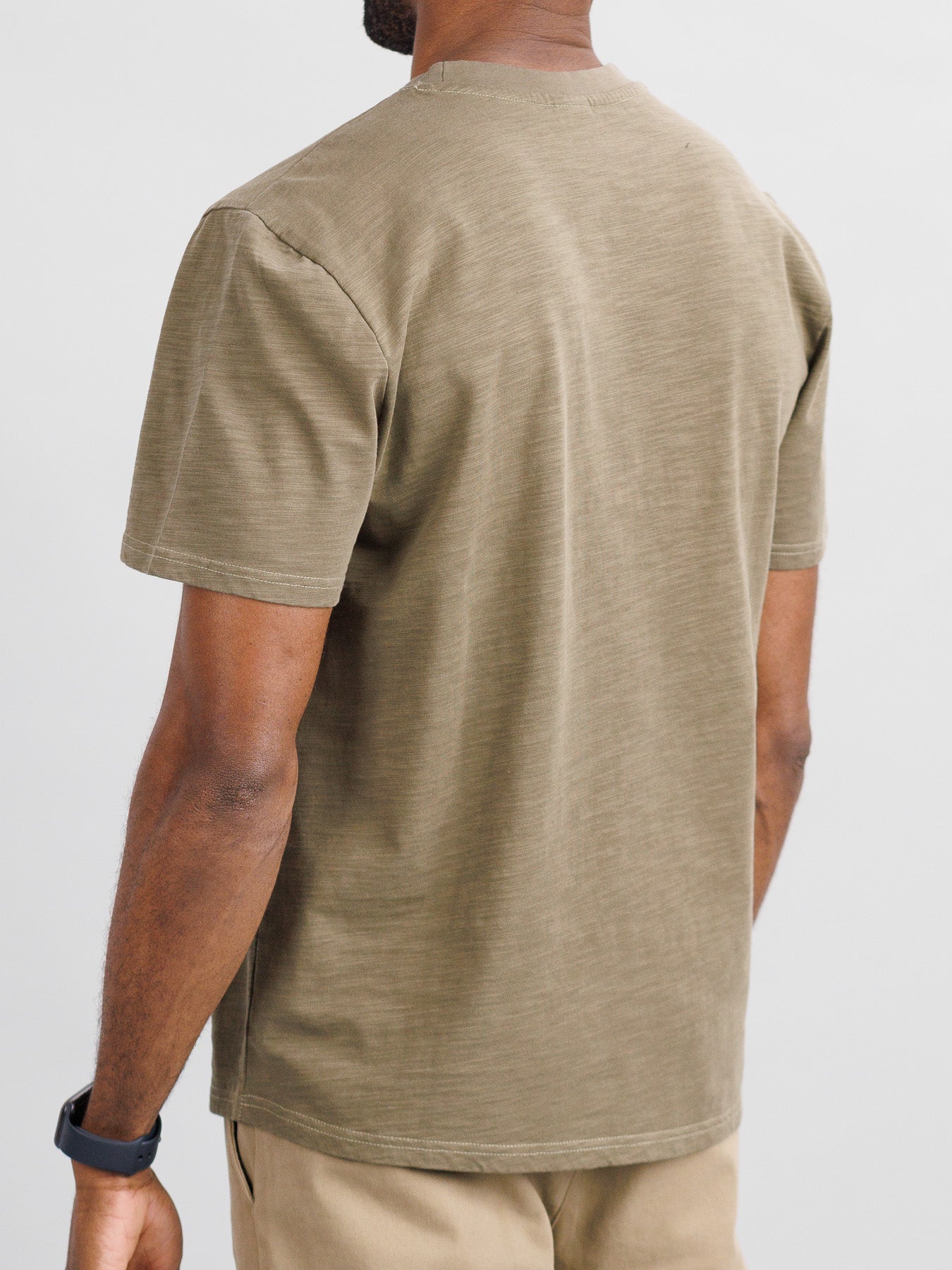 A person wearing ID Menswears Big Flip Slub Yarn Cotton Tee in a boxy fit, paired with plain brown and beige pants, stands back-facing the camera against a white background. The T-shirt drapes casually, while a smartwatch is on their left wrist.