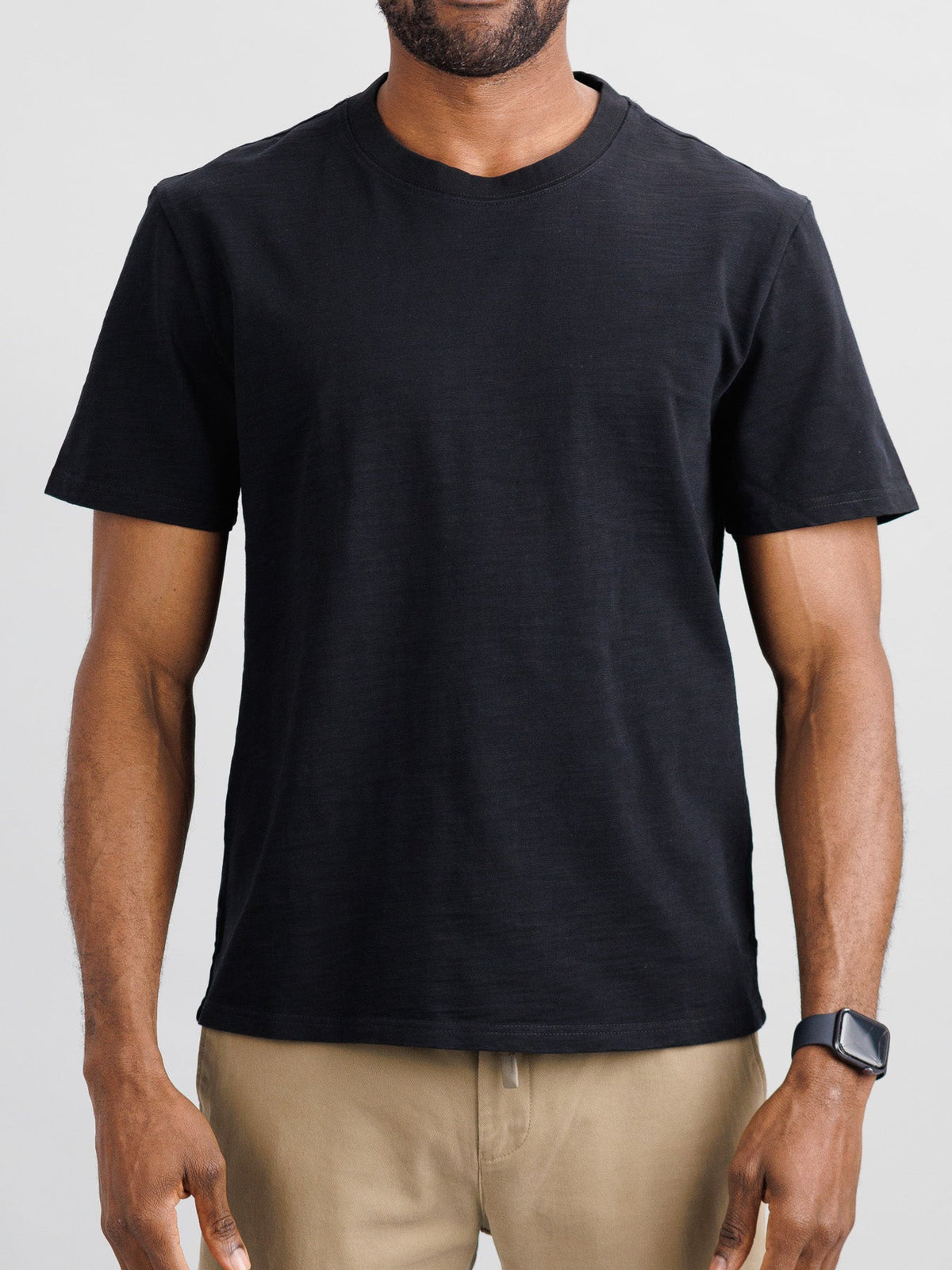 A man in an ID Menswear Big Flip Slub Yarn Cotton Tee and beige pants stands against a neutral background. He sports a boxy fit look, complemented by a black smartwatch on his left wrist.