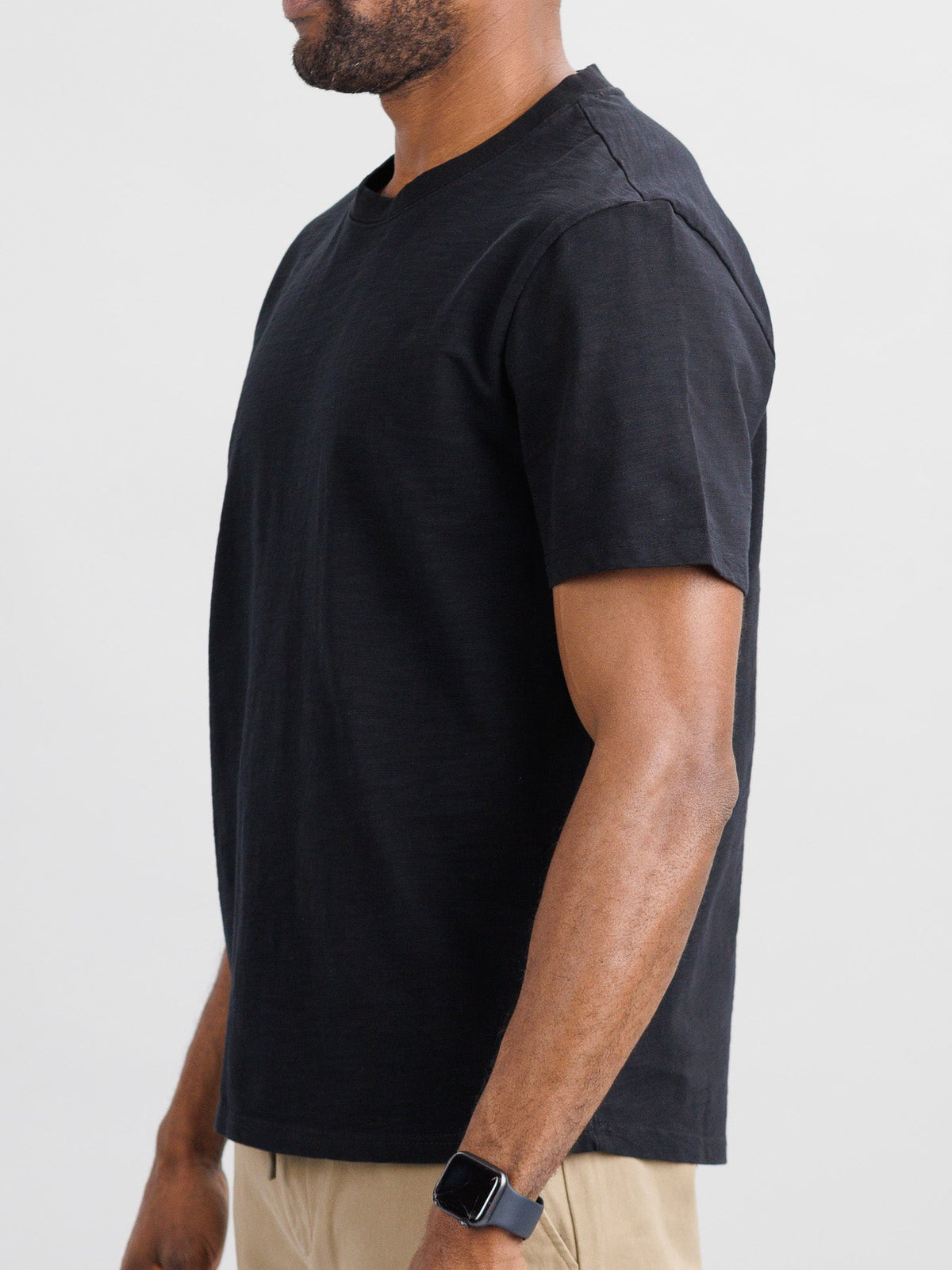 A man in a boxy fit Big Flip Slub Yarn Cotton Tee by ID Menswear, wearing tan pants and a smartwatch, stands against a plain background, shown from the side with arms relaxed.