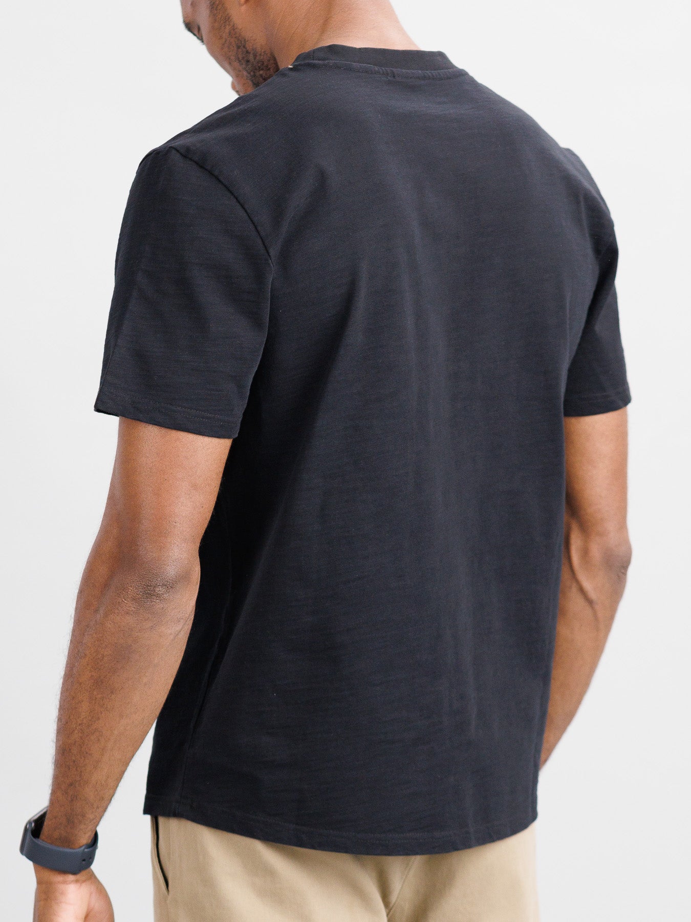 Facing away from the camera, a person wears ID Menswears black Big Flip Slub Yarn Cotton Tee with a boxy fit and beige pants. A smartwatch adorns their left wrist, against a plain, light-colored background.