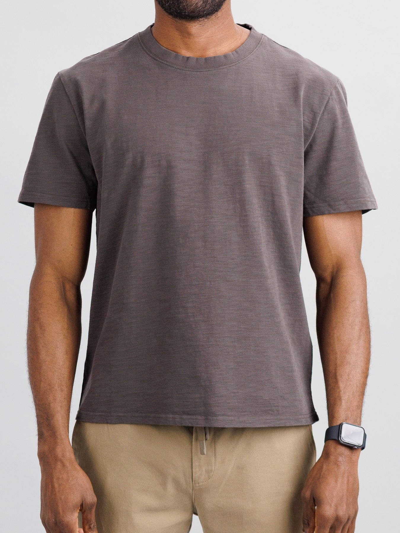 A person wearing the Big Flip Slub Yarn Cotton Tee by ID Menswear, in plain gray, stands against a light background in beige pants. The t-shirt is crafted from 100% cotton with a boxy fit. Their torso and arms are visible along with a smartwatch on their left wrist.