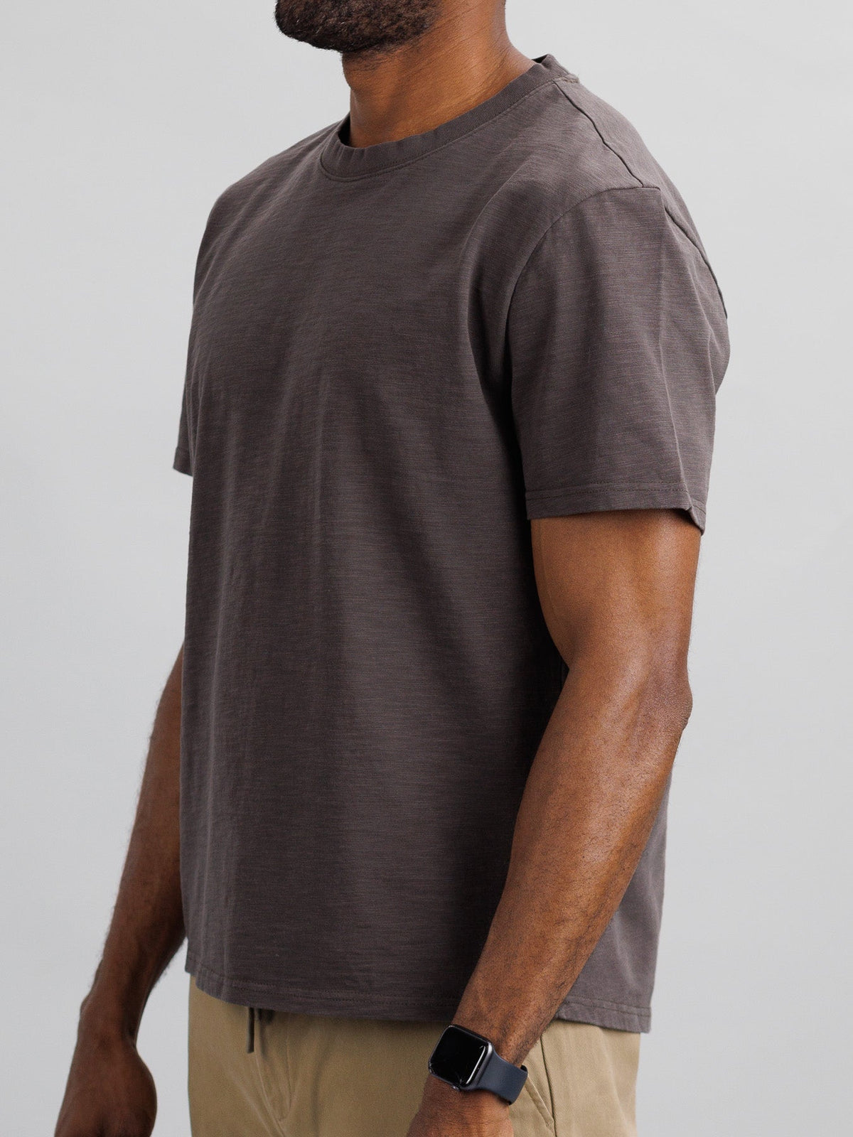 A person, shown from the side against a light gray background, is wearing ID Menswear’s Big Flip Slub Yarn Cotton Tee in plain brown with beige pants. They have dark skin and a smartwatch on their left wrist.
