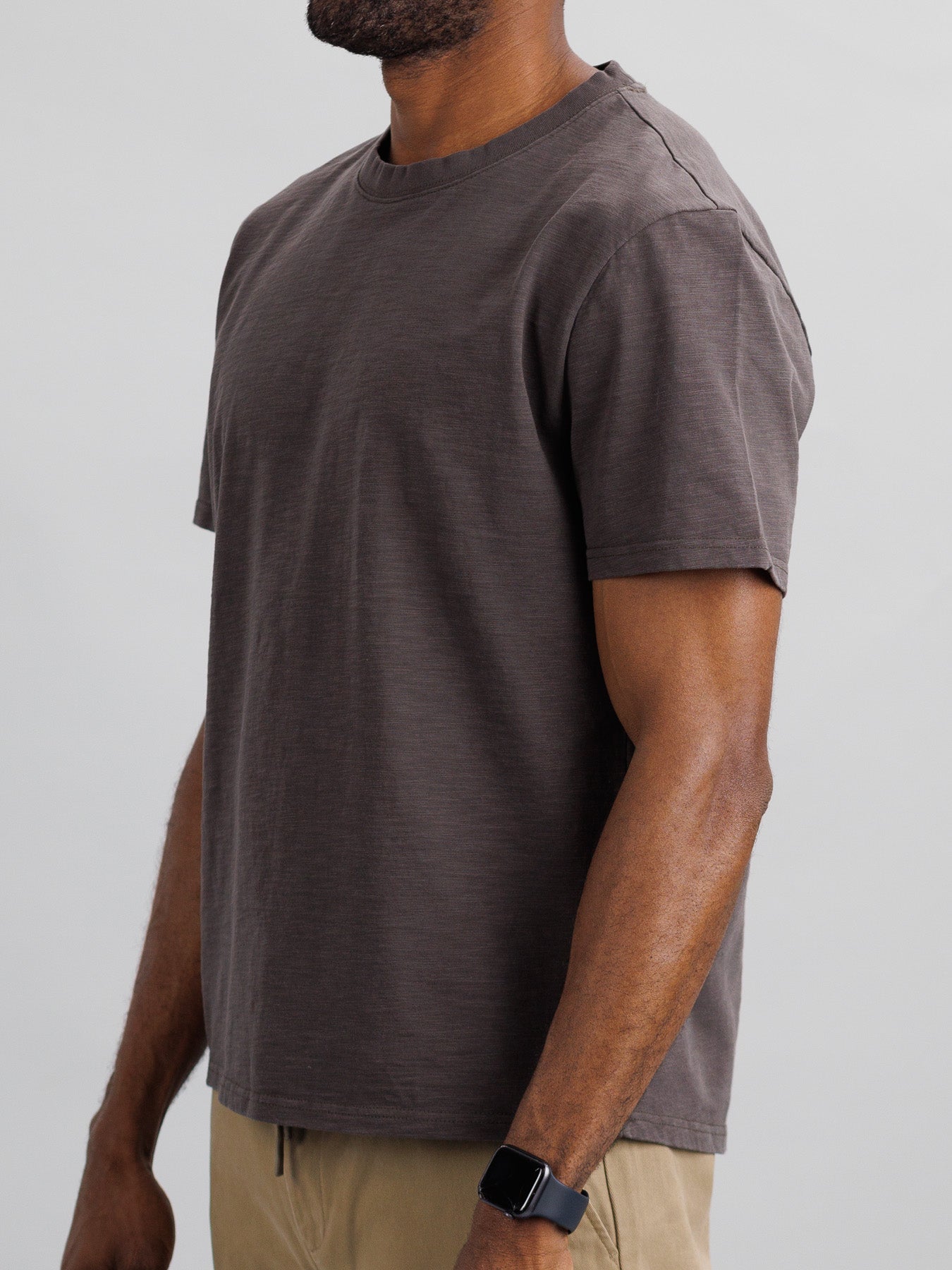 A person, shown from the side against a light gray background, is wearing ID Menswear’s Big Flip Slub Yarn Cotton Tee in plain brown with beige pants. They have dark skin and a smartwatch on their left wrist.