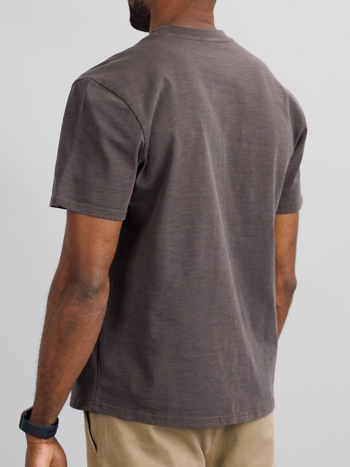 Wearing a dark gray Big Flip Slub Yarn Cotton Tee by ID Menswear and beige pants, a person stands with their back to the camera. The T-shirt pairs well with the smartwatch on their left wrist against a plain light gray background.