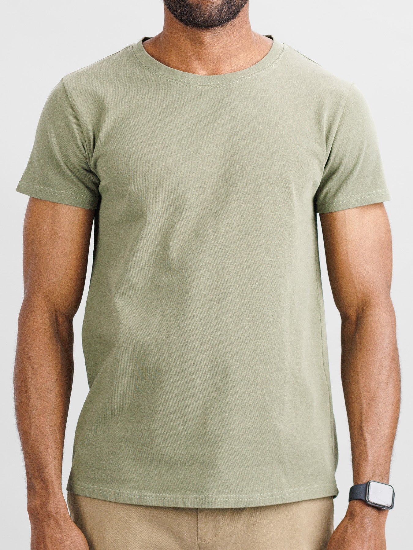 A man dressed in an ID Menswear City Organic Cotton Tee, featuring light green and slim-fit design, paired with beige pants stands against a neutral background. He sports short hair, an unshaven face, and a smartwatch on his right wrist.
