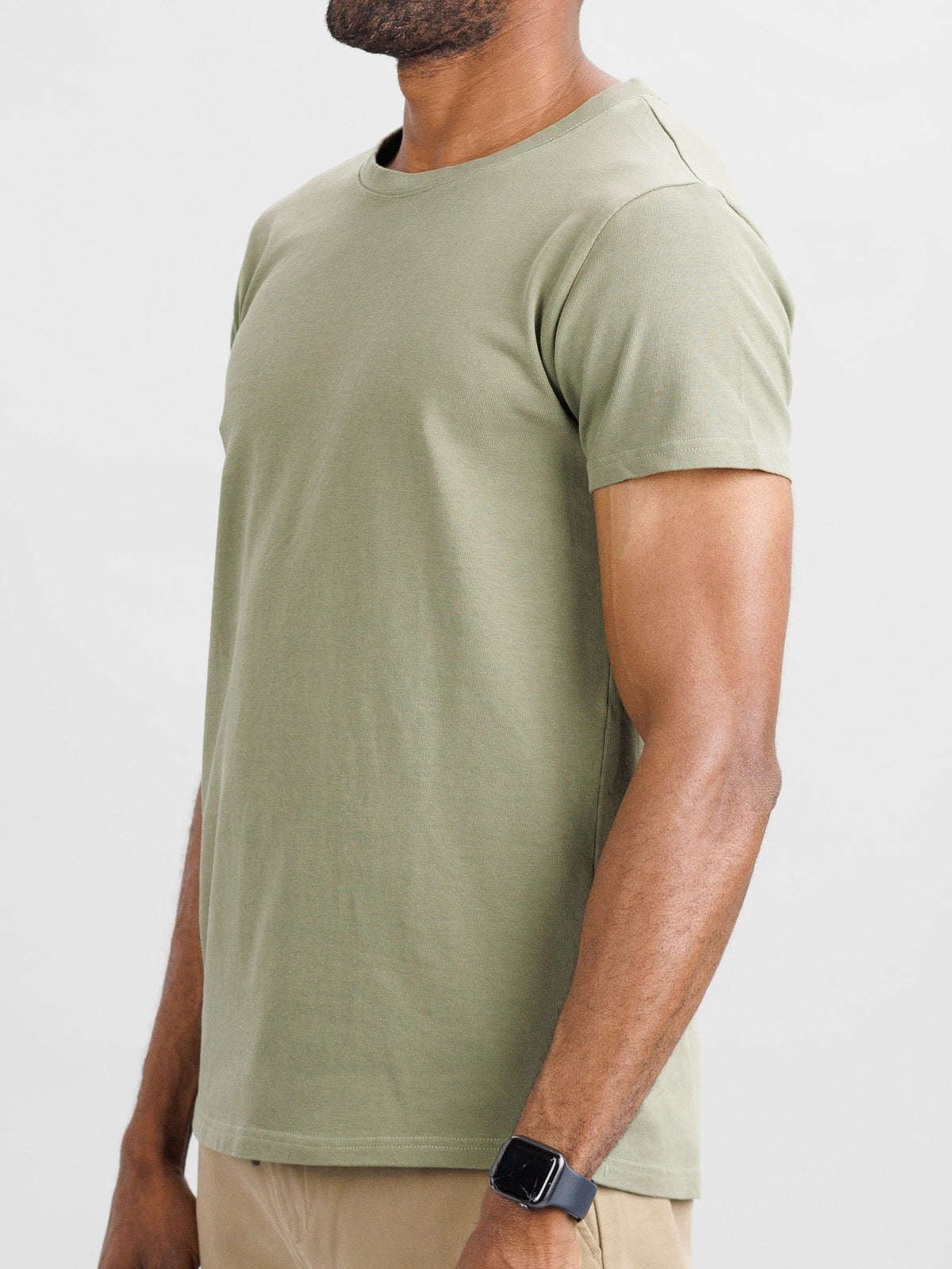 A person in a City Organic Cotton Tee by ID Menswear, slim fit in light green, with beige pants, stands sideways. The tee matches the smartwatch on their wrist against a plain white backdrop.