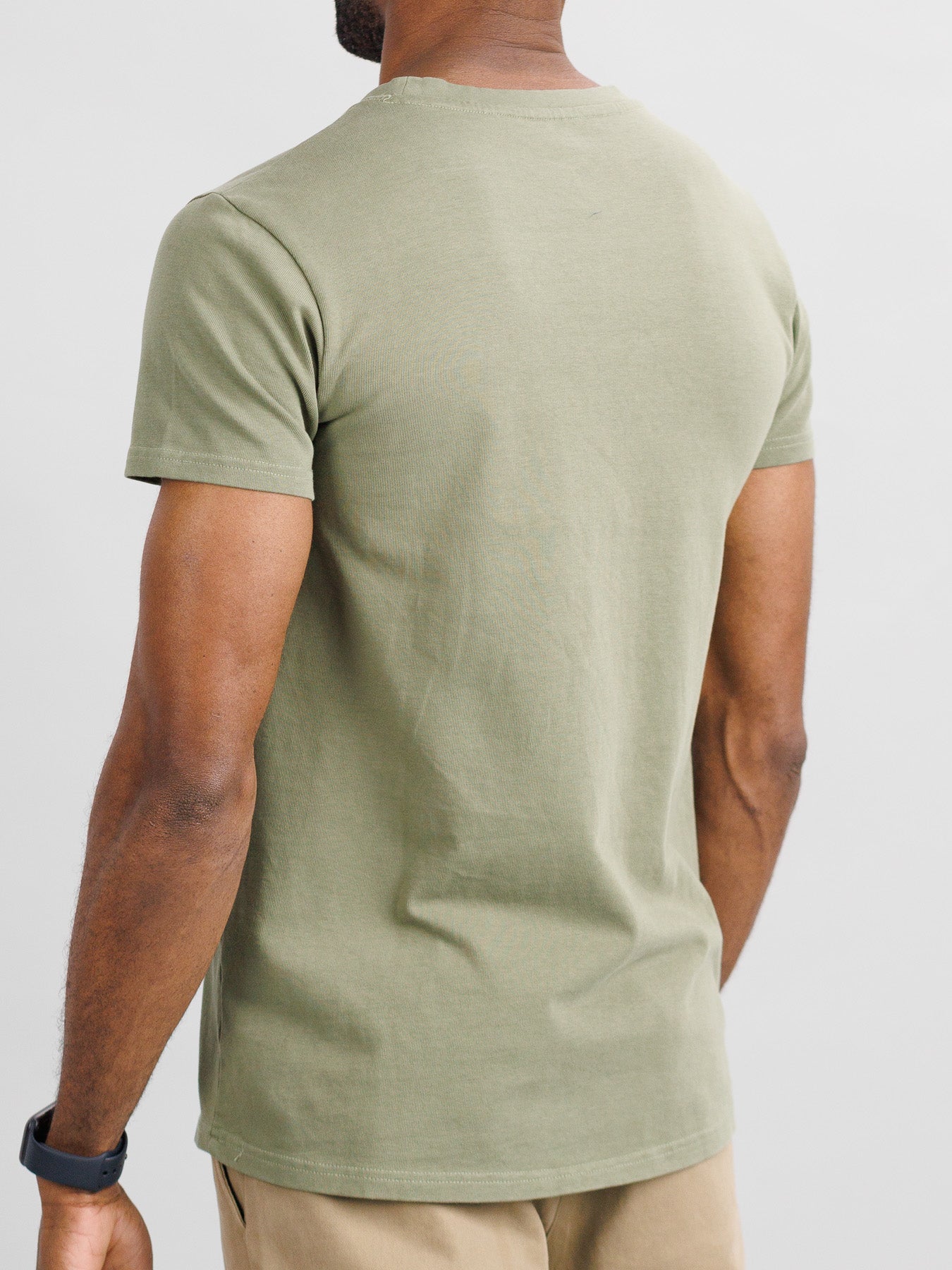 A person wearing the ID Menswear City Organic Cotton Tee in olive green and beige pants stands with their back turned, highlighting the slim fit tee. A smartwatch adorns their left wrist against a light gray backdrop.