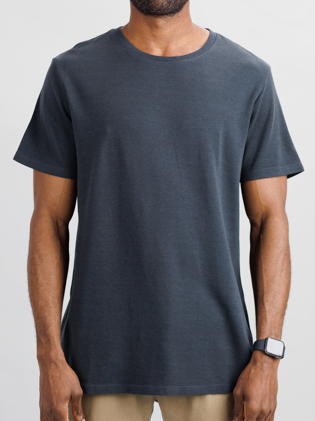 A person in an Urban Honeycomb Mesh Tee by ID Menswear, dark gray and made from a breathable bamboo-organic cotton blend, paired with beige pants, stands against a neutral background. Theyre wearing a smart watch on their left wrist, though their face is not visible.