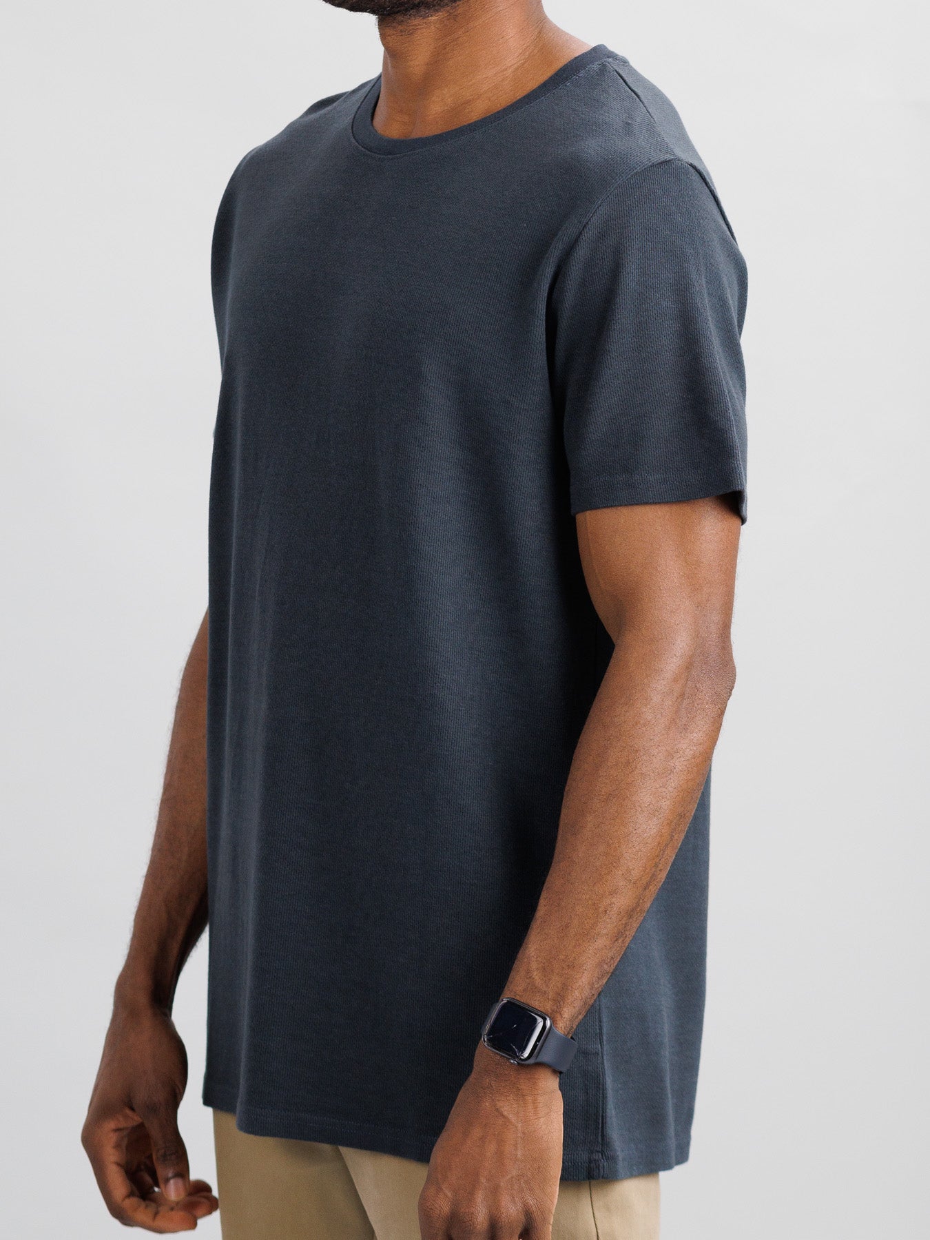 A man wearing an Urban Honeycomb Mesh Tee from ID Menswear, in dark gray, crafted from a breathable bamboo-organic cotton blend, stands with his smartwatch visible and arms resting at his sides against a light background.