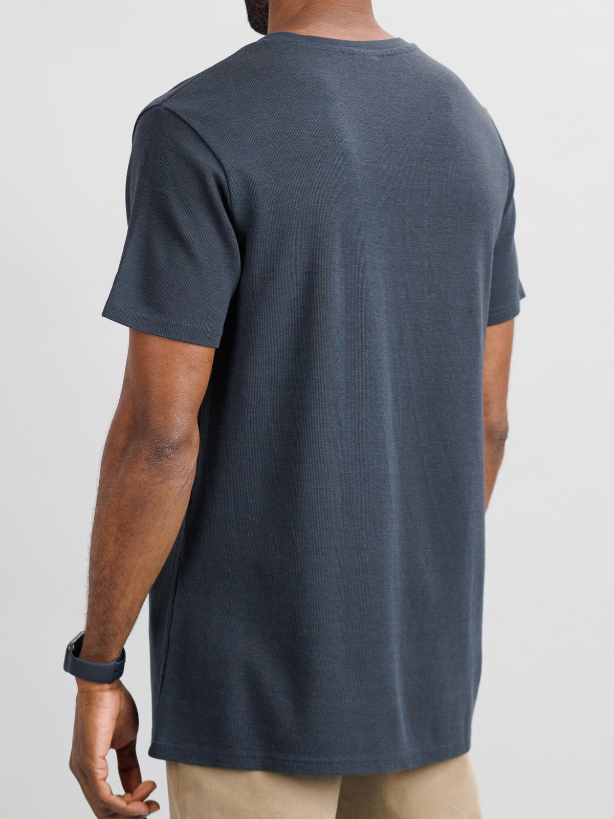 A person is seen from the rear, wearing ID Menswears Urban Honeycomb Mesh Tee in dark gray paired with beige pants. They have short hair and a black wristwatch, set against a light gray background that highlights the T-shirts breathable bamboo-organic cotton blend texture.