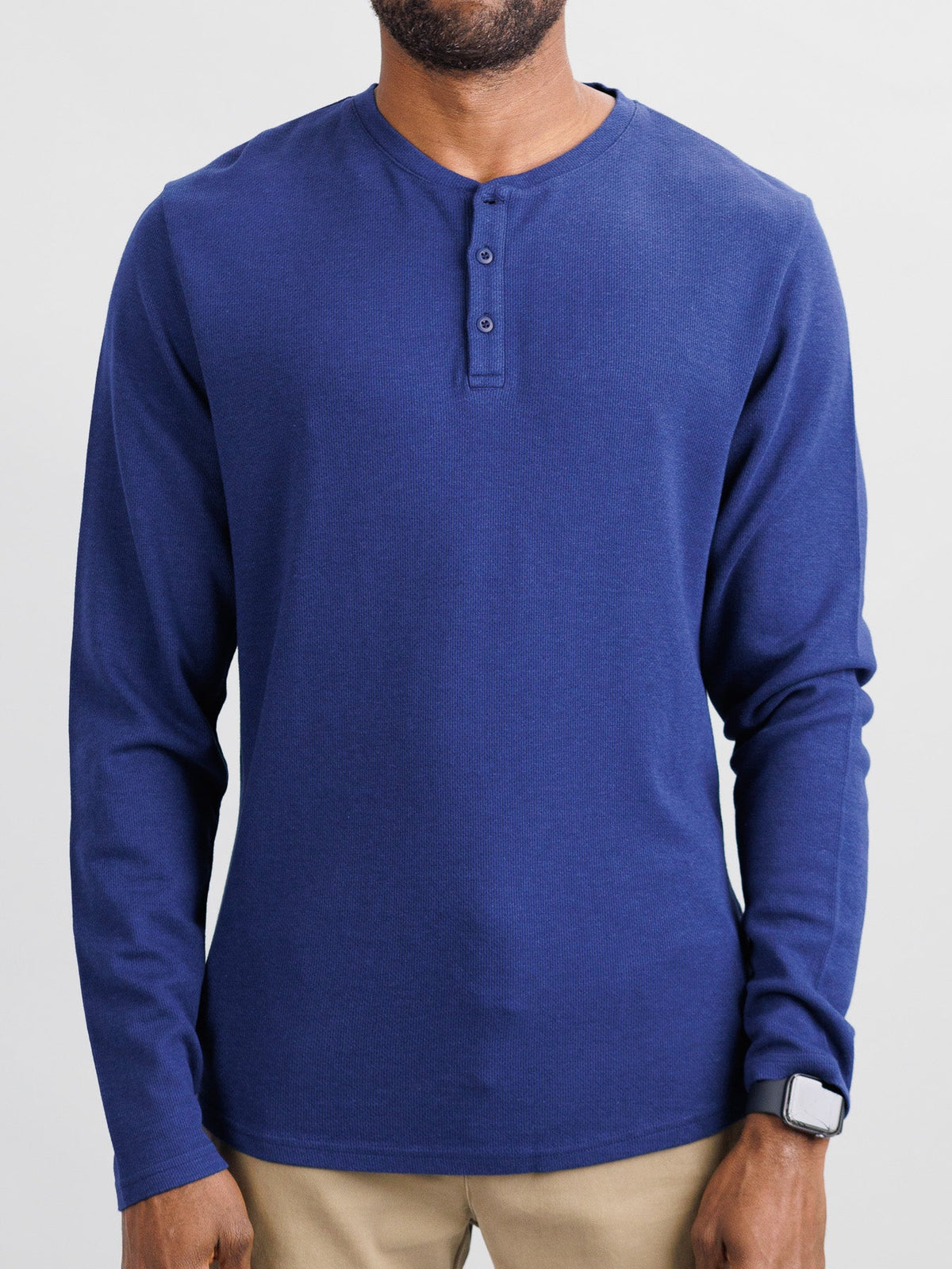 A person in ID Menswears Freestyle Honeycomb Knitted Mesh Henley, a long-sleeve blue organic cotton shirt with a buttoned placket, stands facing forward on a white background. They wear a smartwatch on their left wrist and light beige pants.