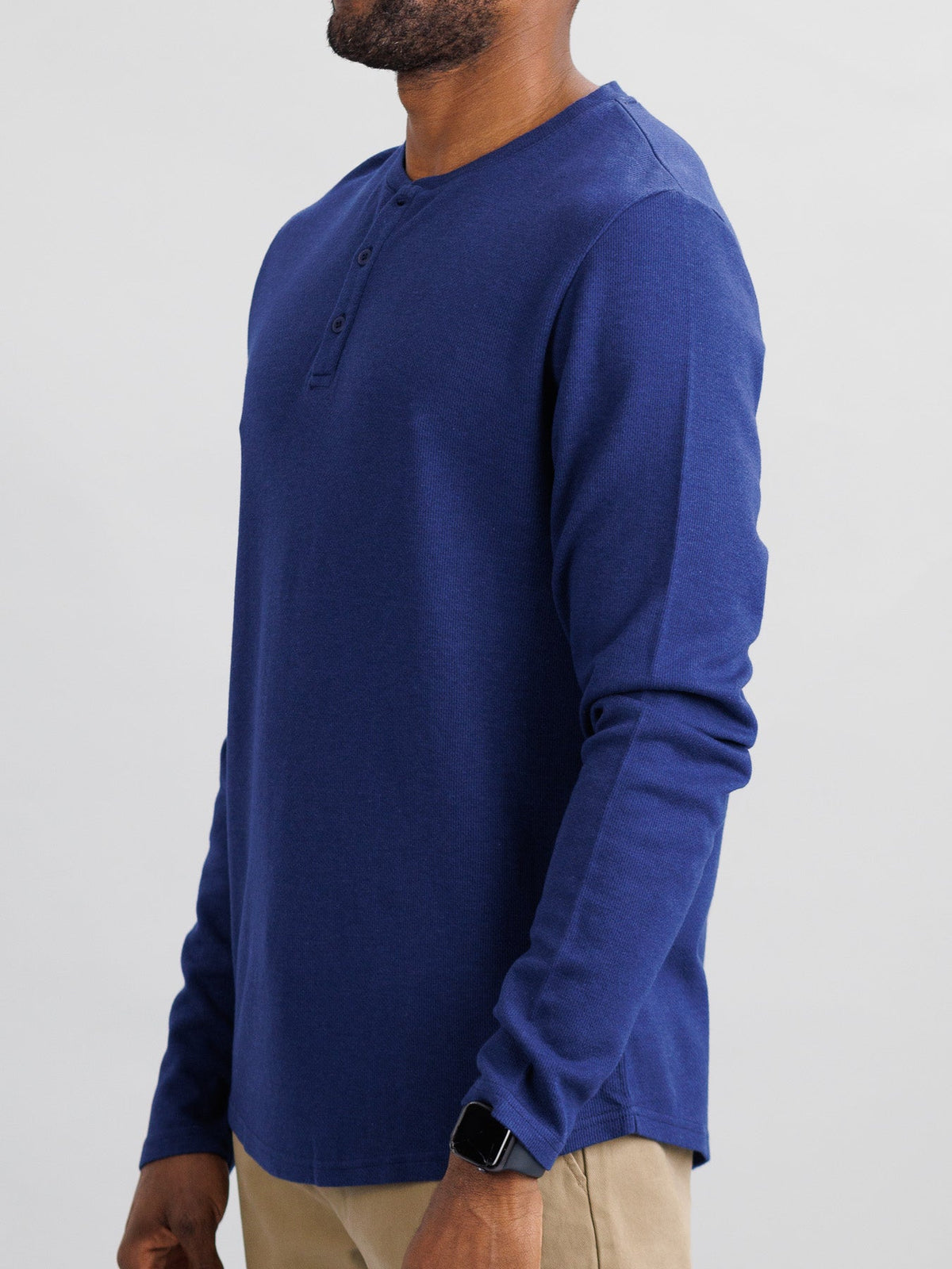 A person stands sideways, arms relaxed, dressed in ID Menswears Freestyle Honeycomb Knitted Mesh Henley and beige pants, wearing a smartwatch on their wrist against a plain light-colored background.