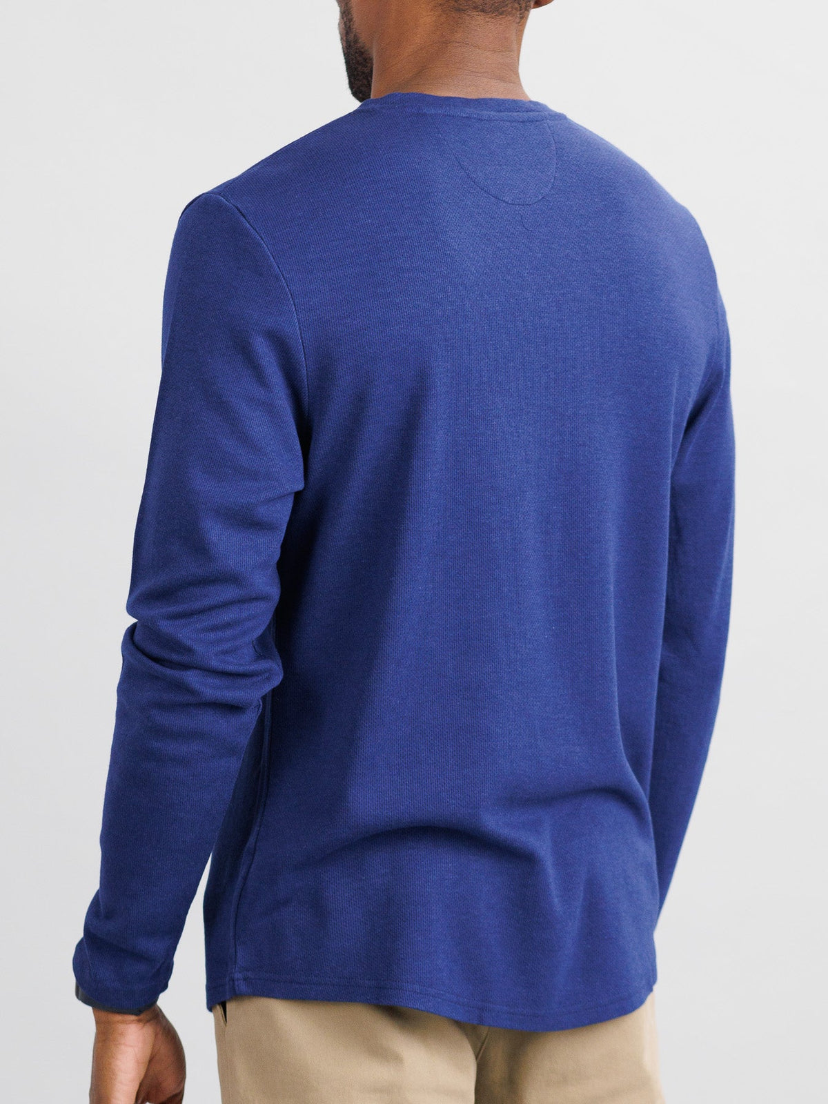 A person wearing the Freestyle Honeycomb Knitted Mesh Henley from ID Menswear, a blue organic cotton long-sleeve shirt, and beige pants stands with their back to the camera against a plain background.