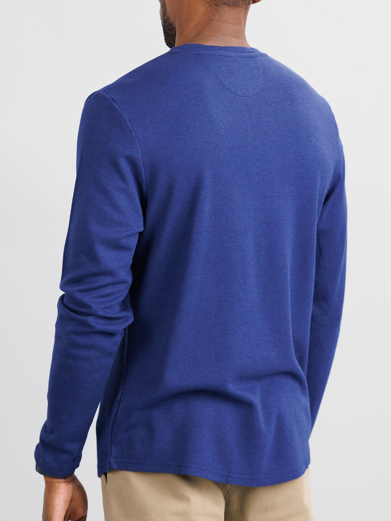 A person wearing the Freestyle Honeycomb Knitted Mesh Henley from ID Menswear, a blue organic cotton long-sleeve shirt, and beige pants stands with their back to the camera against a plain background.