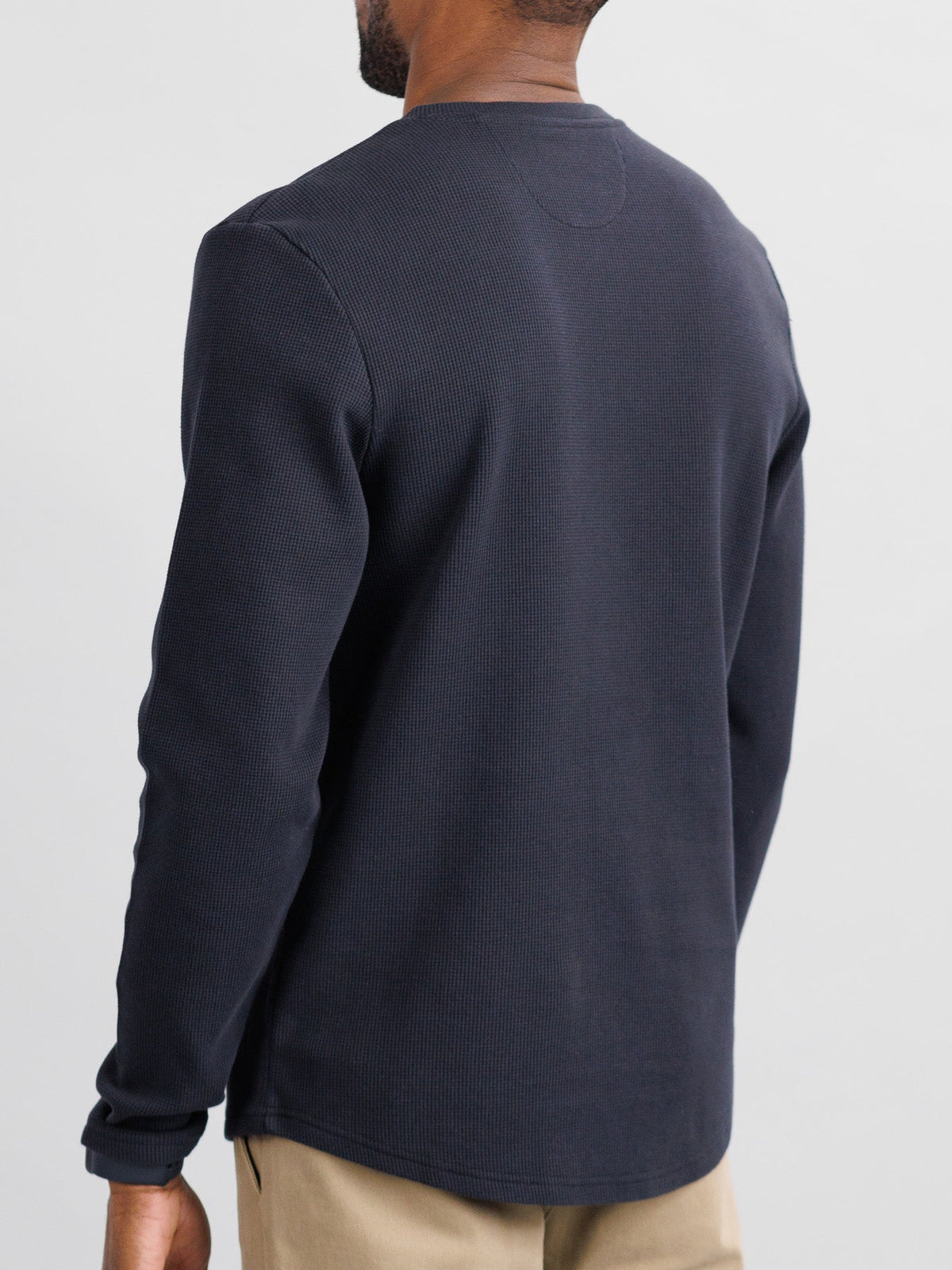 A person wearing an ID Menswear Freestyle Honeycomb Knitted Mesh Henley in long sleeves and black cotton, stands facing away. Their short hair pairs well with light-colored pants against a plain light gray wall.