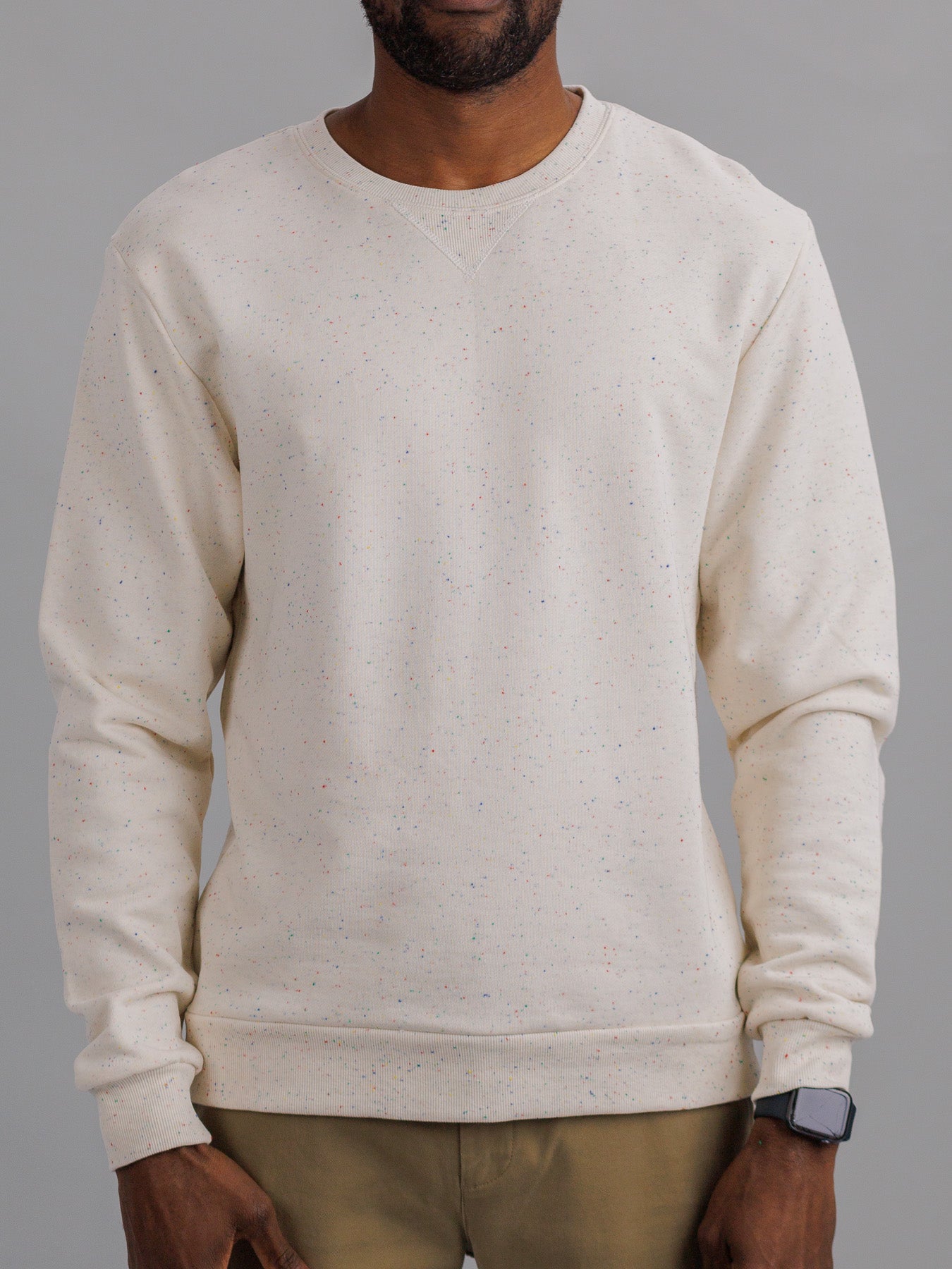 Against a gray background, an individual in ID Menswears cream-colored Stardust Speckled Sweatshirt and khaki pants exudes casual style. The regular fit cotton sweatshirt pairs seamlessly with a smartwatch on the left wrist, blending fashion and technology effortlessly.