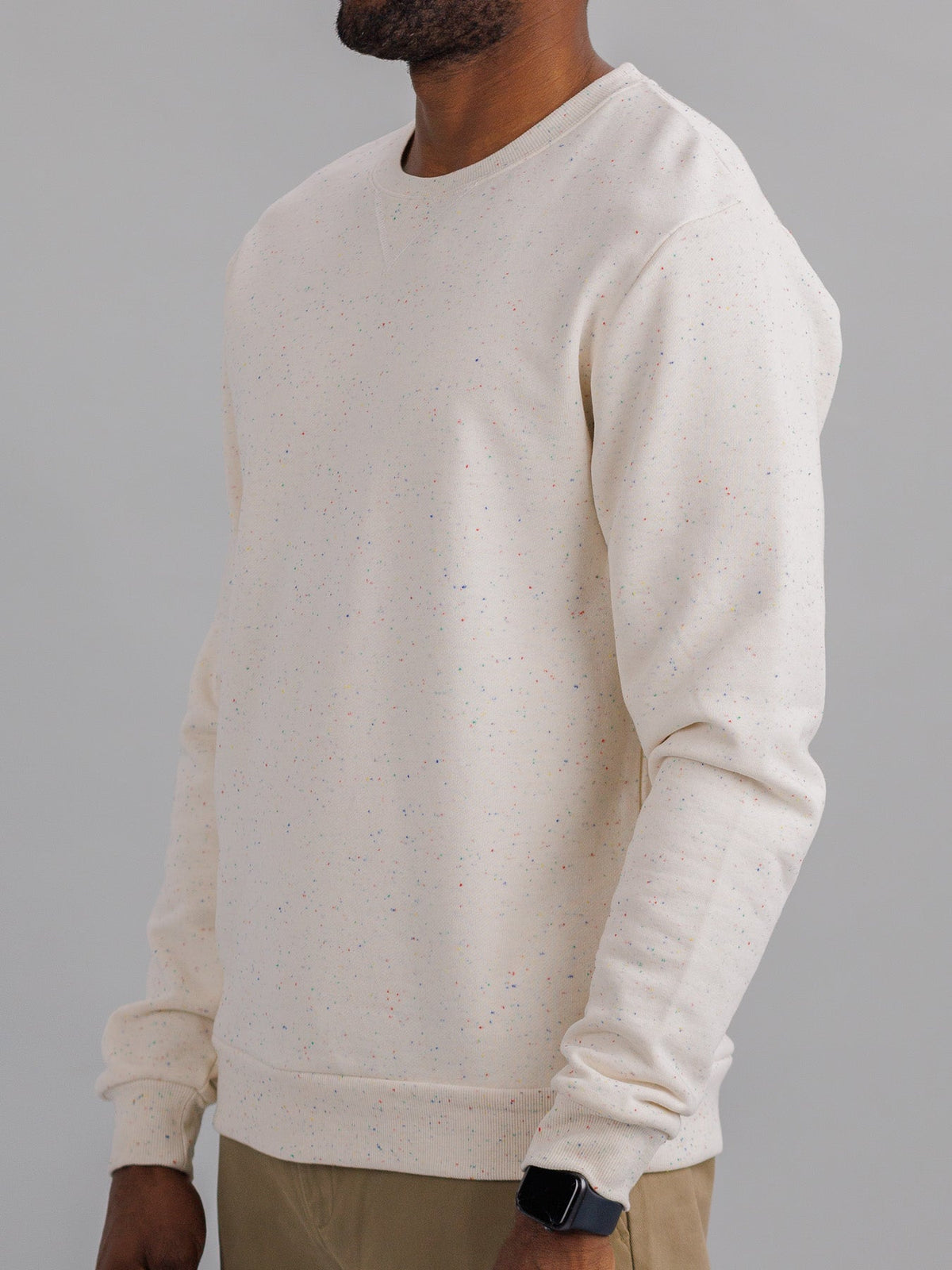 A person in an ID Menswear Stardust Speckled Sweatshirt, crafted from light beige cotton yarn, stands sideways to display their upper body. Paired with khaki pants and a smartwatch, the plain gray background enhances the subtle vibrancy of their attire.
