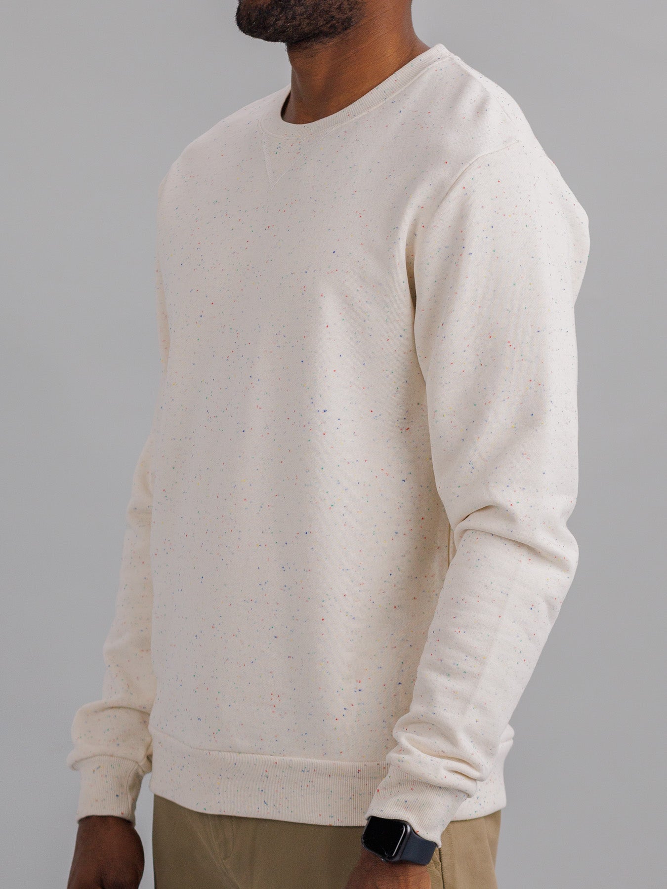 A person in an ID Menswear Stardust Speckled Sweatshirt, crafted from light beige cotton yarn, stands sideways to display their upper body. Paired with khaki pants and a smartwatch, the plain gray background enhances the subtle vibrancy of their attire.