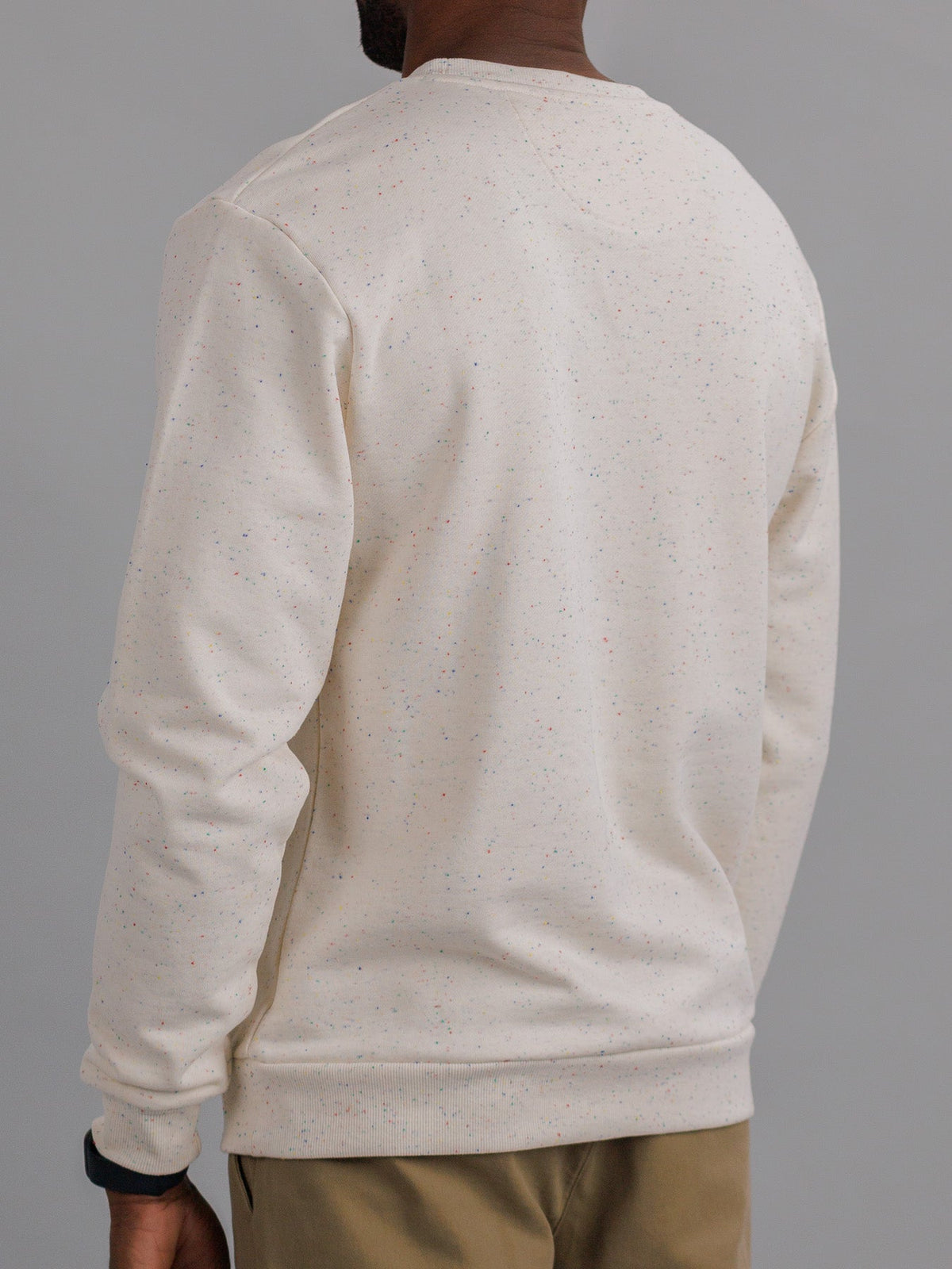 Wearing ID Menswears Stardust Speckled Sweatshirt in light beige, paired with light brown pants, the person is shown from the back against a gray backdrop. Crafted from speckled cotton yarn.