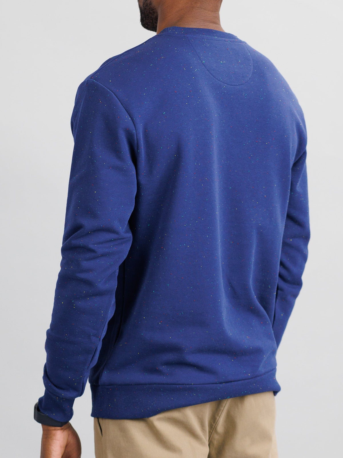 A person in an ID Menswear Stardust Speckled Sweatshirt and beige pants stands against a plain light background with short hair, wearing a dark watch on their left wrist.