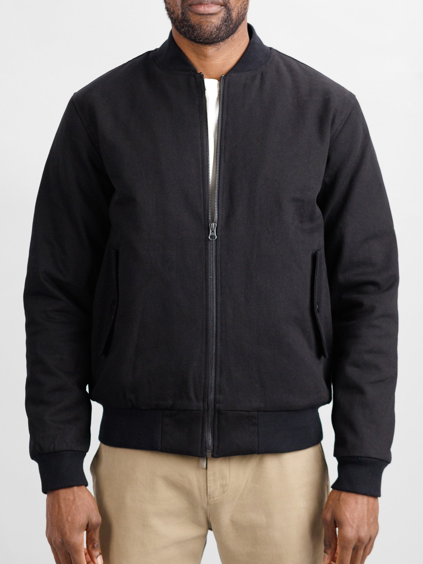 Someone is wearing the Yonkers Cotton Canvas Jacket from ID Menswear, a black bomber with quilted lining, ribbed collar, cuffs, hem, and side pockets. Its styled over a white shirt and beige pants against a plain light-colored background.