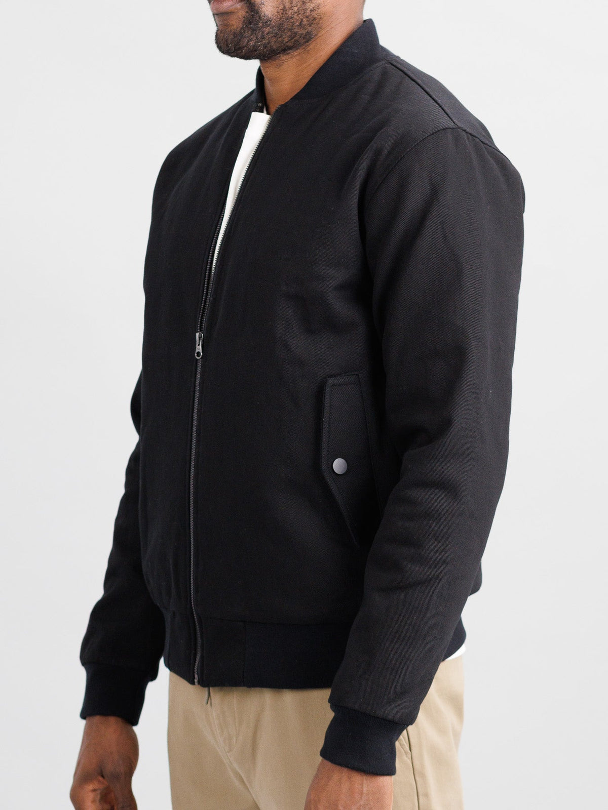 A person is wearing the Yonkers Cotton Canvas Jacket by ID Menswear, a black bomber with quilted lining, over a white shirt and beige pants. The cotton-padded jacket includes a front zipper and side buttoned pockets. They are posed neutrally against a plain background.