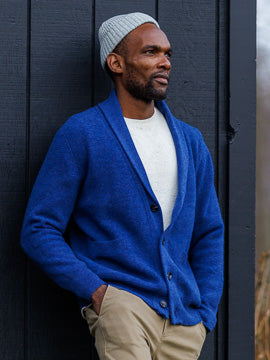 A man in a grey beanie, ID Menswears Basecamp Cashmere Merino Cardigan in blue, a white shirt, and beige pants leans against a dark wooden wall, gazing to the side.
