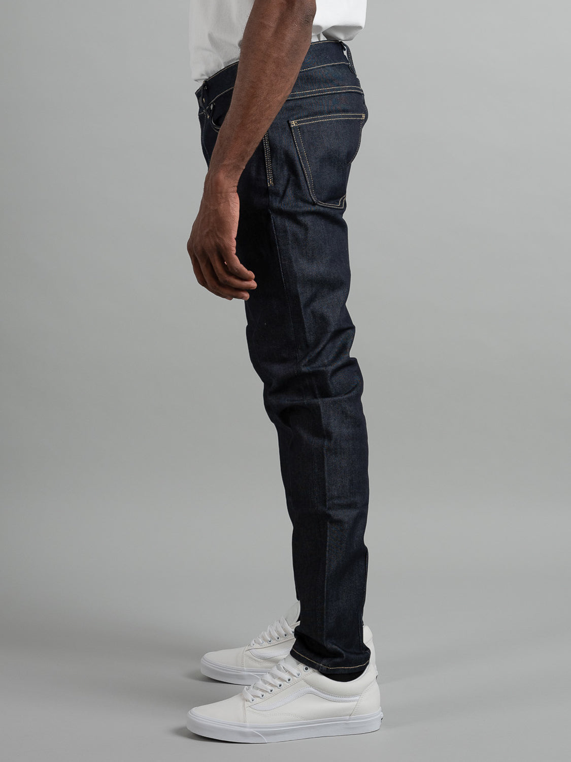 A person stands sideways wearing Neuw Lou Slim Raw Stretch jeans, a white t-shirt, and white sneakers against a plain gray background.