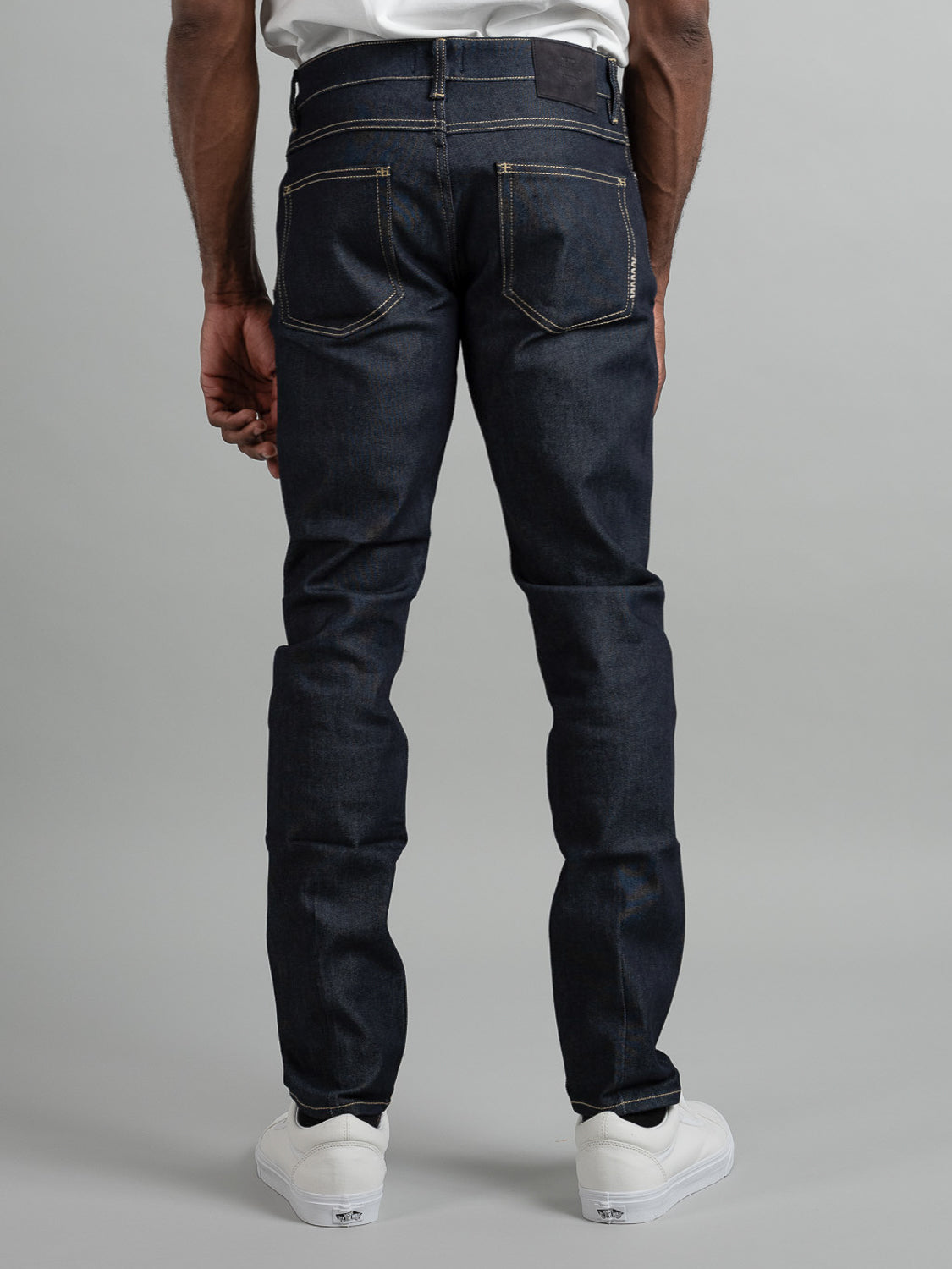 A person stands facing away against a plain gray background, wearing Neuw Lou Slim Raw Stretch jeans with yellow stitching and white sneakers.