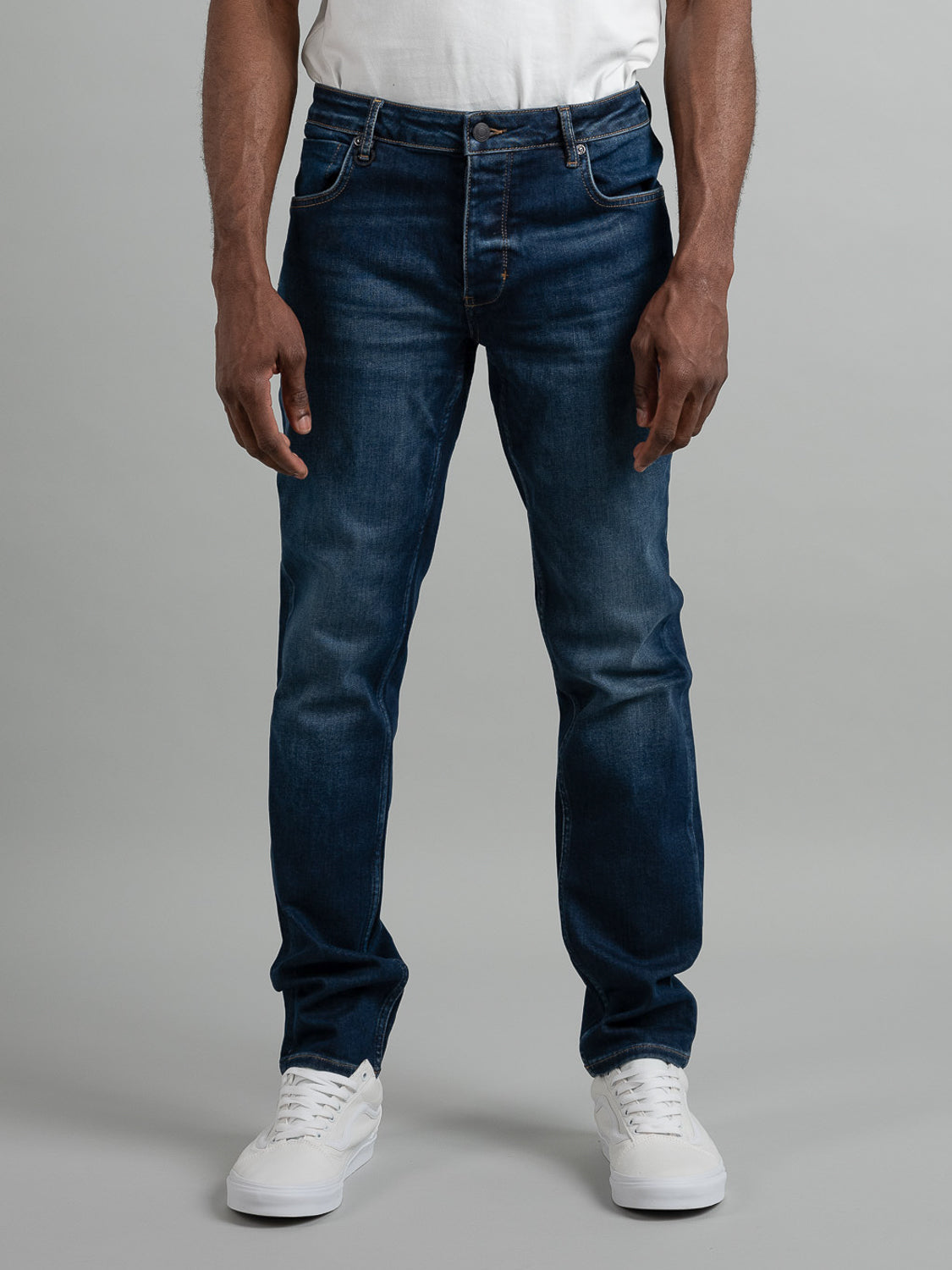 A person wearing a white T-shirt, Lou Slim Blue Sunday jeans by Neuw, and white sneakers stands facing forward against a plain gray background, showing only their lower torso and legs.