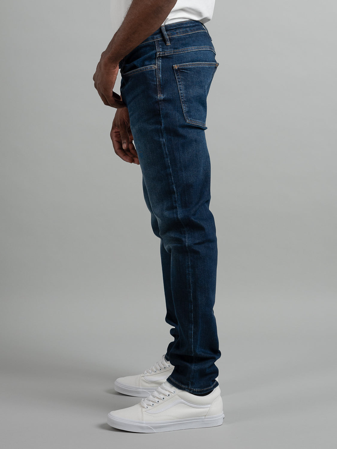A person stands sideways against a plain gray background, showing only their lower body in Neuw Lou Slim Blue Sunday jeans and white sneakers, from waist to feet.