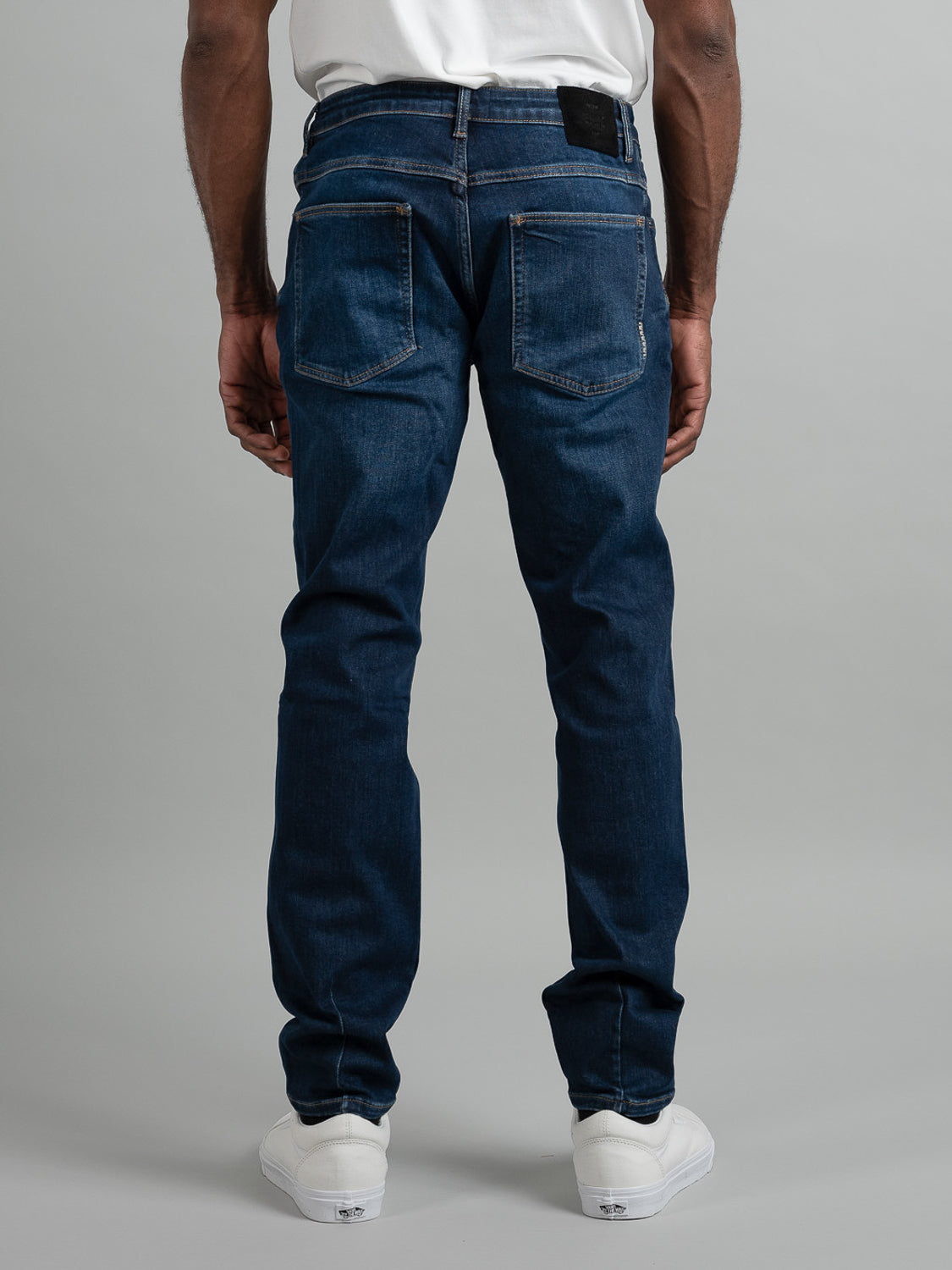 Someone is wearing Neuw Lou Slim Blue Sunday jeans, a white t-shirt, and white sneakers, standing with their back to the camera against a plain gray background.