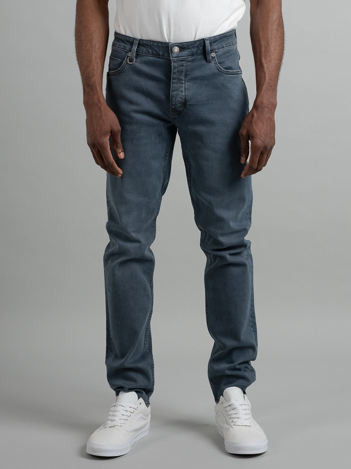 A person stands against a gray background, wearing the Neuw Lou Slim Mercury faded blue slim leg jeans, a white t-shirt, and white sneakers. The photo is cropped at the shoulders to highlight the jeans and shoes.