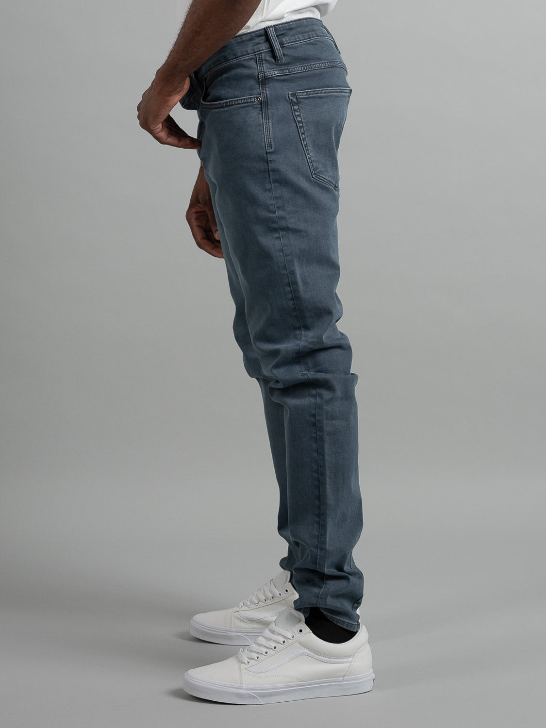 A person stands side-on against a plain gray background wearing Neuw Lou Slim Mercury jeans, a white t-shirt, black socks, and white sneakers.